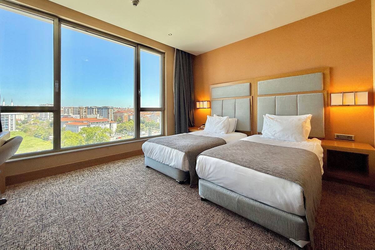 Ein Hotelzimmer mit zwei Betten, einem großen Fenster mit Blick auf die Stadt, einem Schreibtisch und einem Nachttisch mit Lampe.