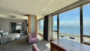 Habitación de hotel con un ventanal grande que ofrece vistas al mar y barcos. Se ve una cama, una silla y una mesa con dos copas de vino.