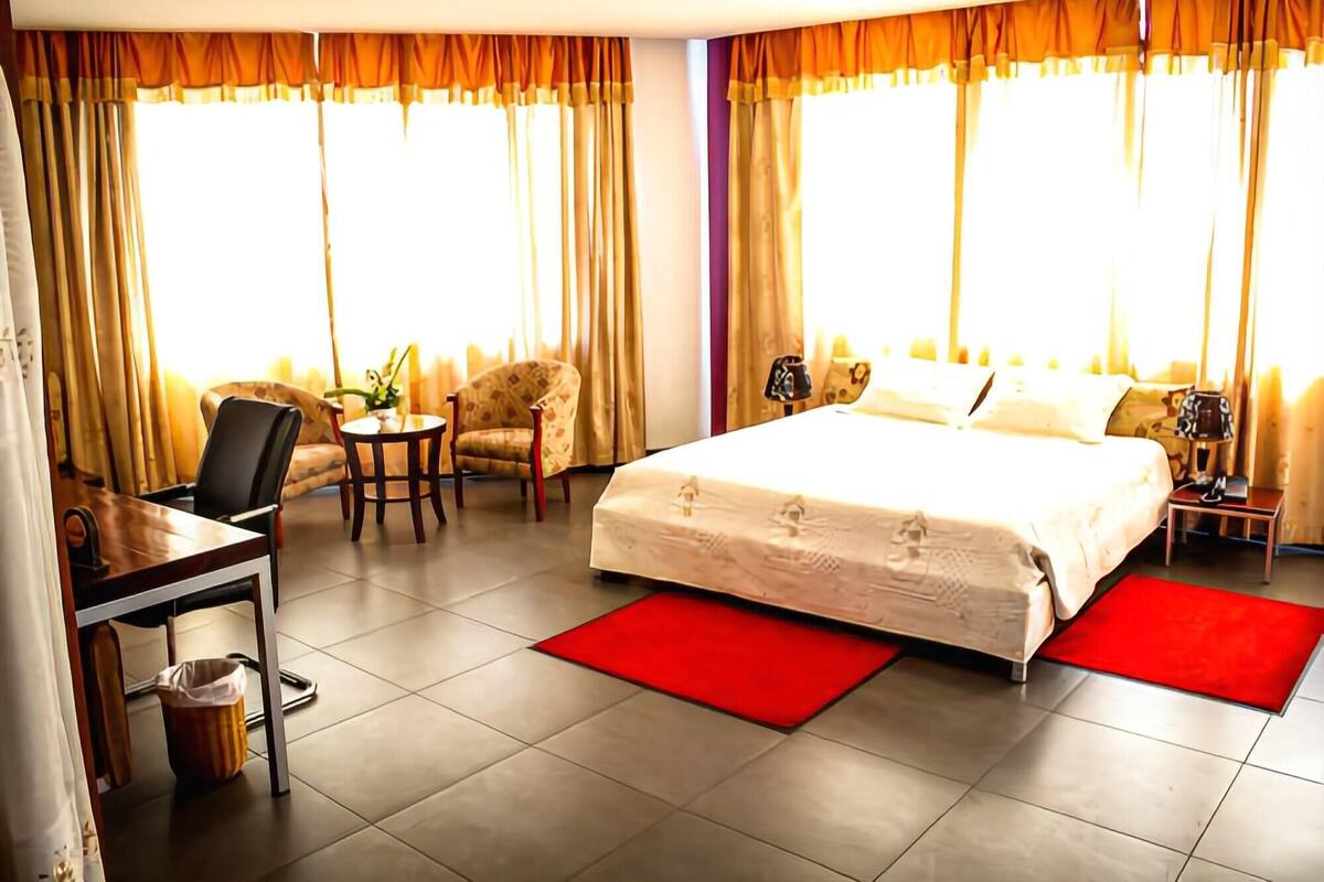 A hotel room with a bed, a desk, a chair, a small table, and curtains.
