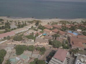 Vista aérea de un complejo turístico frente al mar, con varias construcciones, una playa y palmeras.