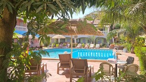 Zona de piscina con sillones de descanso y sombrillas, rodeada de vegetación y edificios.