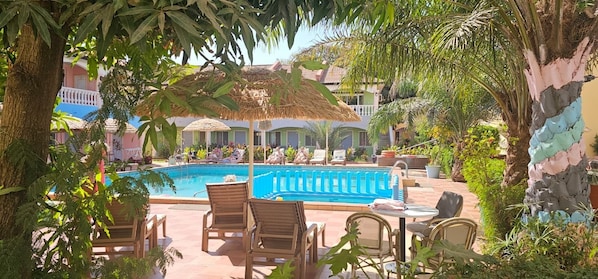 Un espace piscine doté de chaises longues et de parasols, entouré de verdure et de bâtiments.