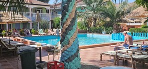 Un área de piscina con sillones reclinables y una mesa, rodeada de palmeras y un edificio con balcón.