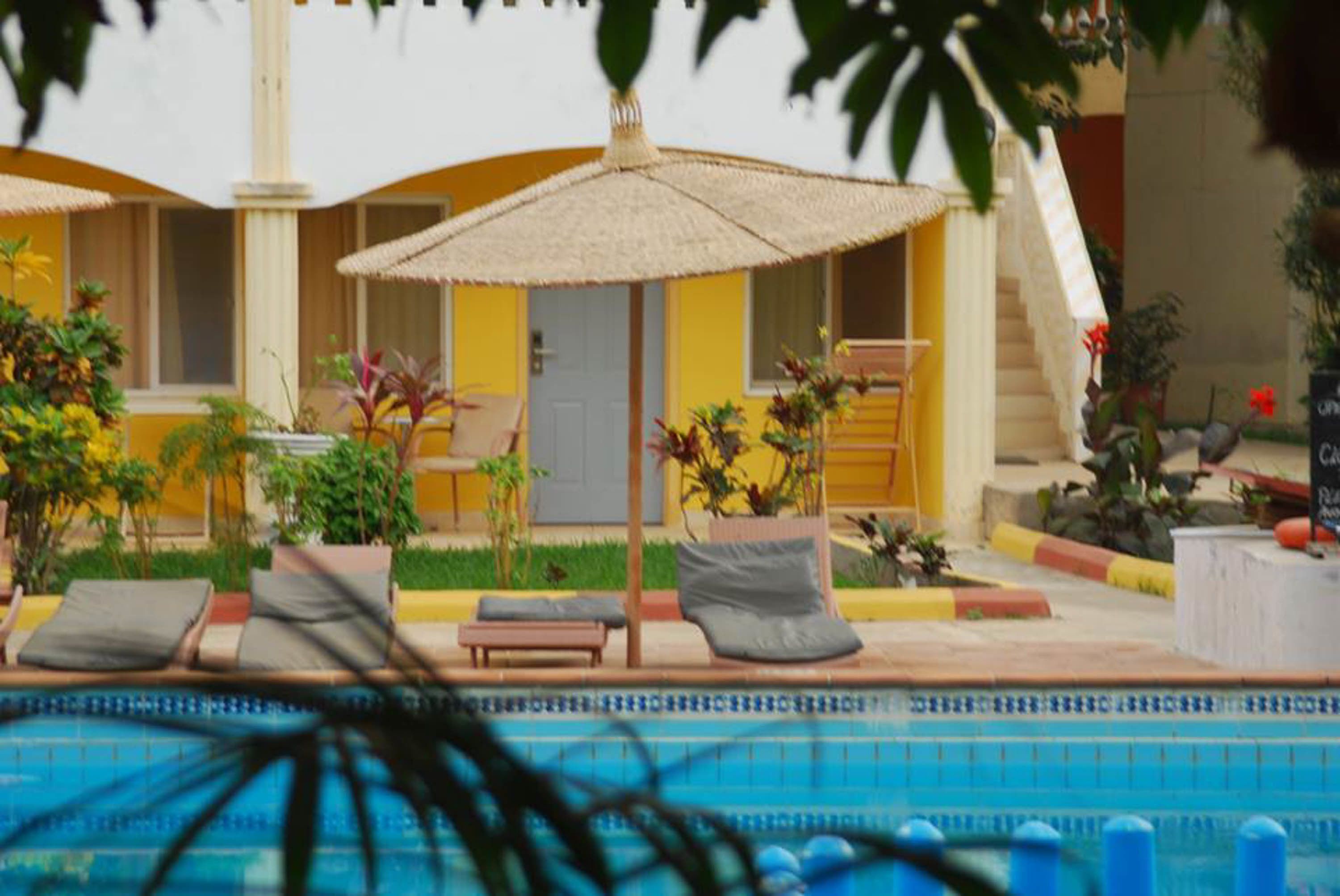 A pool area with lounge chairs and umbrellas, surrounded by potted plants and a building with a yellow facade.