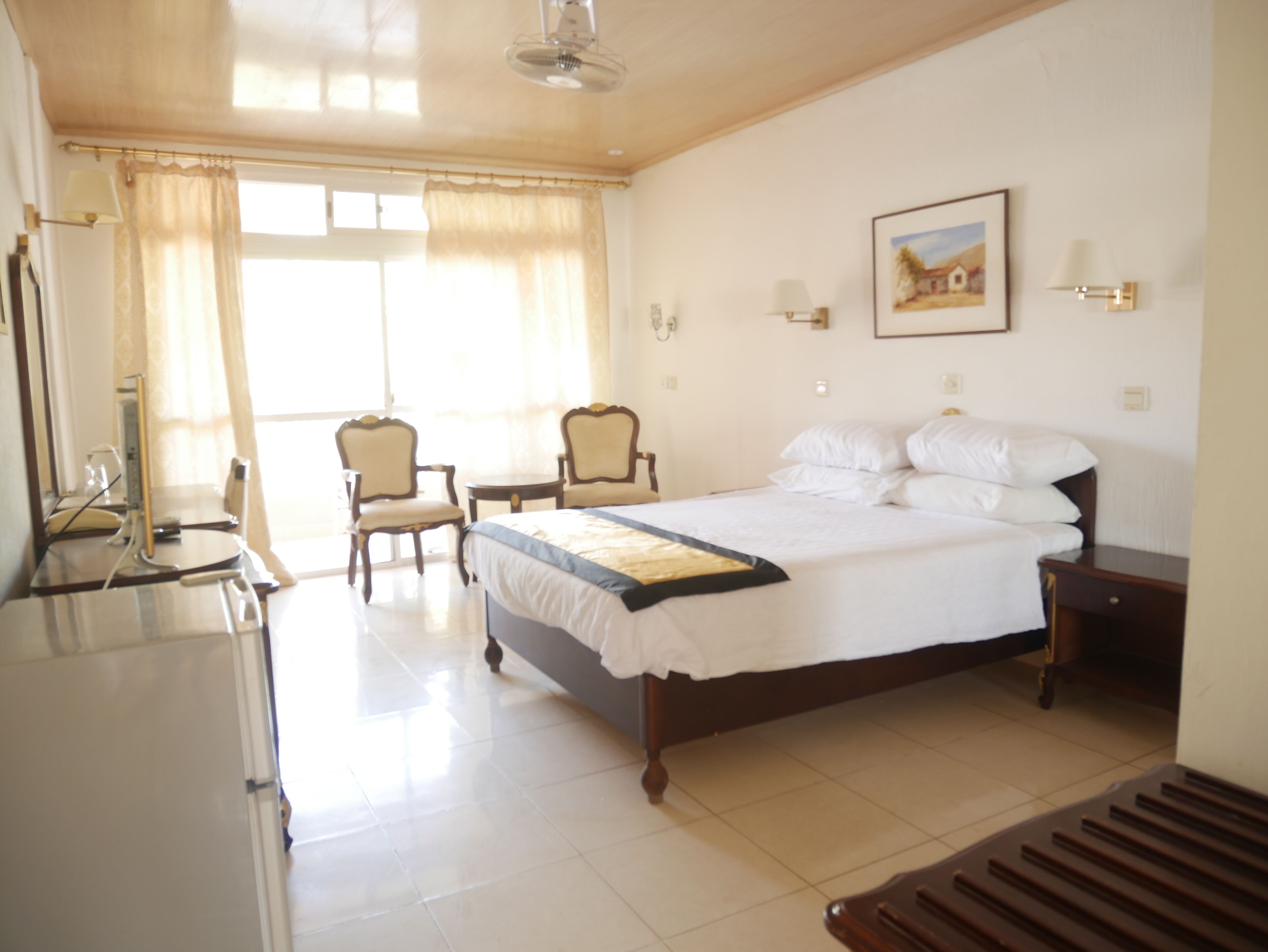 Habitación de hotel con cama, dos sillas, escritorio, lámpara y un cuadro en la pared.