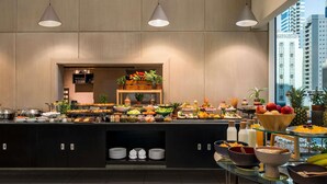 A modern hotel breakfast buffet with a variety of food options, a fruit display, and a view of the cityscape. - ibis Sharq Kuwait (Kuwait City)