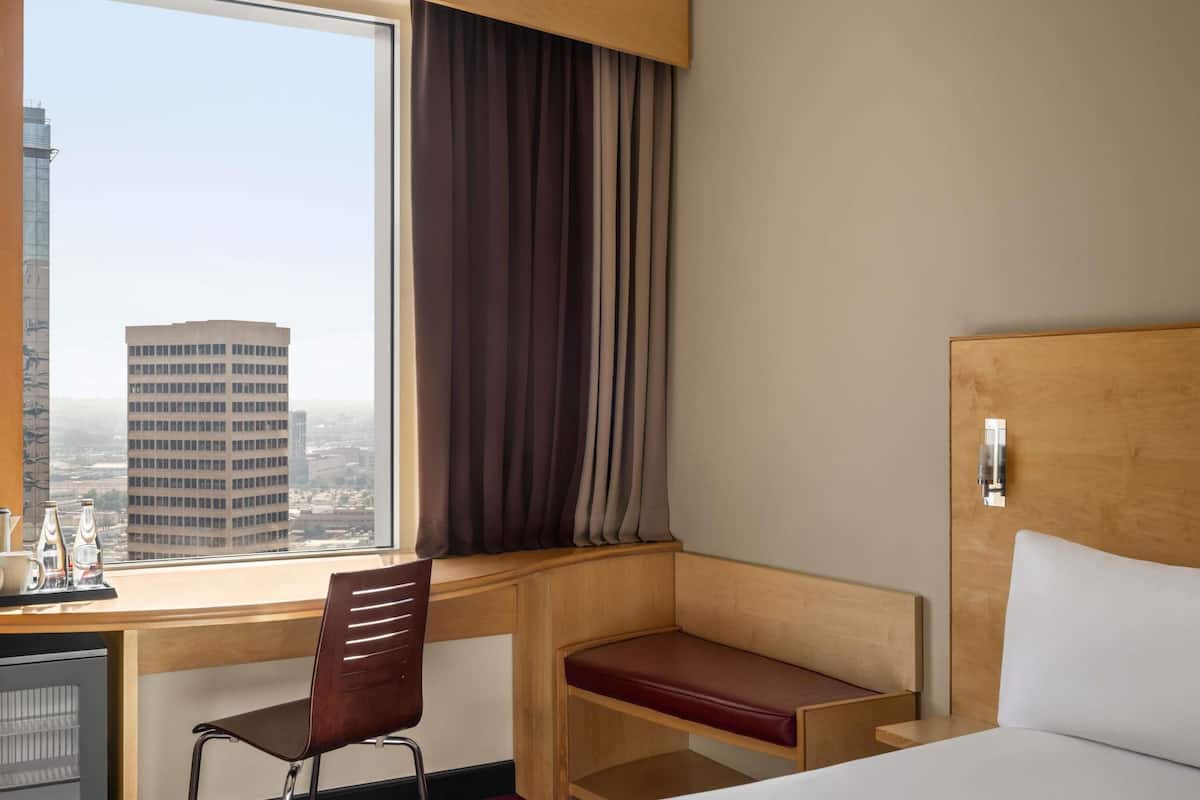 Habitación de hotel con cama, escritorio, silla, ventana con cortinas y vista panorámica de la ciudad.