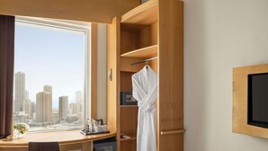A hotel room with a large window, a desk with a mini-fridge, a robe hanging, and a TV mounted on the wall. - ibis Sharq Kuwait (Kuwait City)