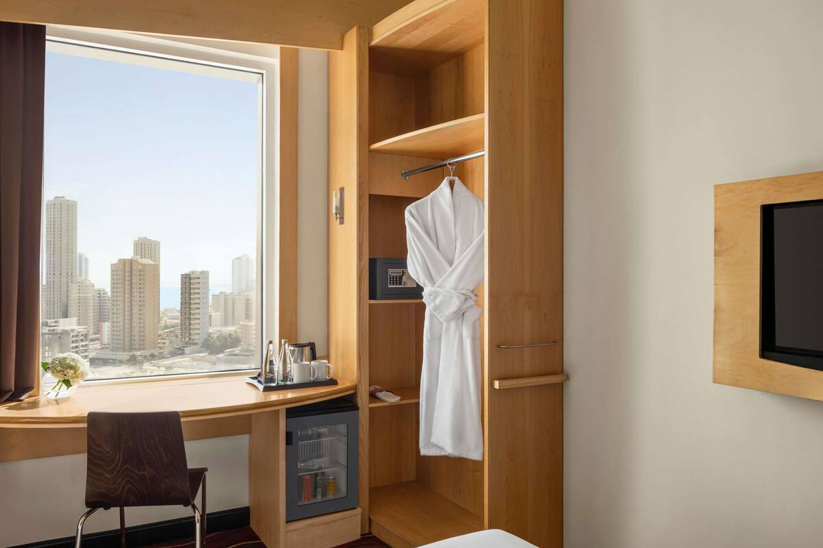 Habitación de hotel con un ventanal grande, un escritorio con mini refrigerador, una bata colgada y un televisor montado en la pared.