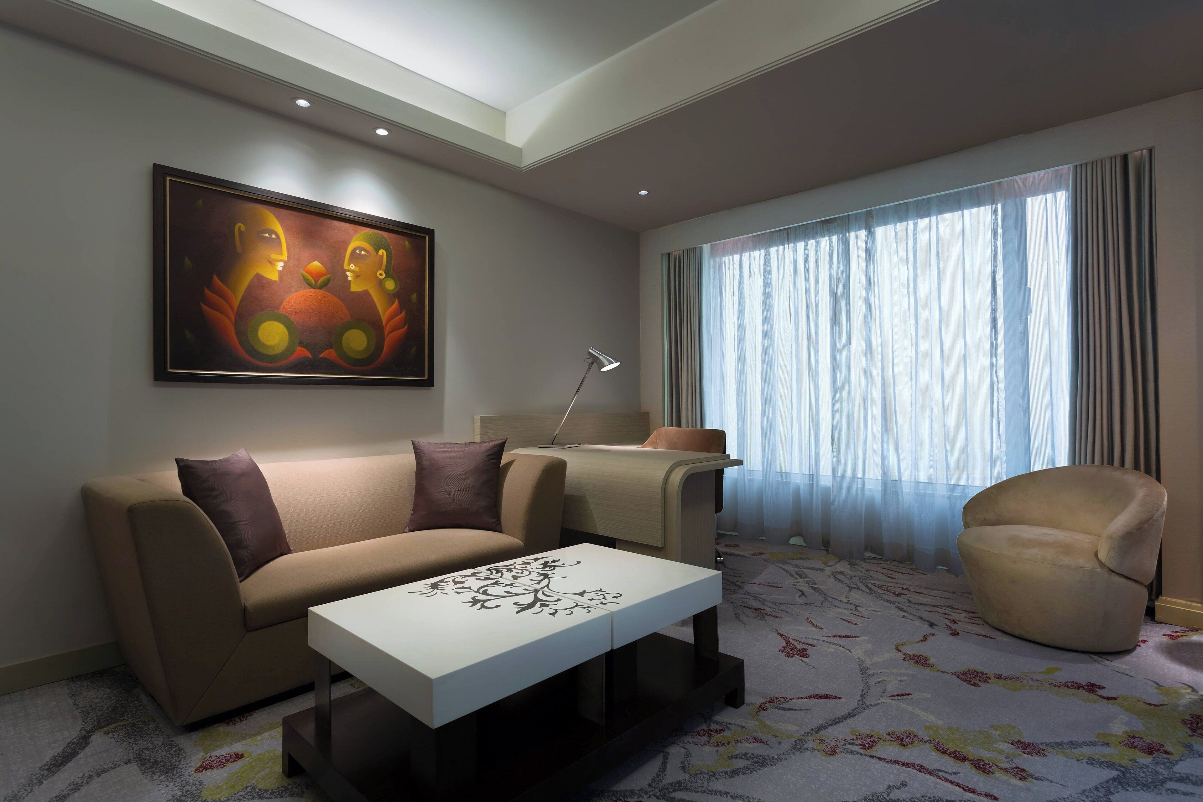A modern living room with a sofa, armchair, and a coffee table. There is a painting on the wall and a lamp on a side table.