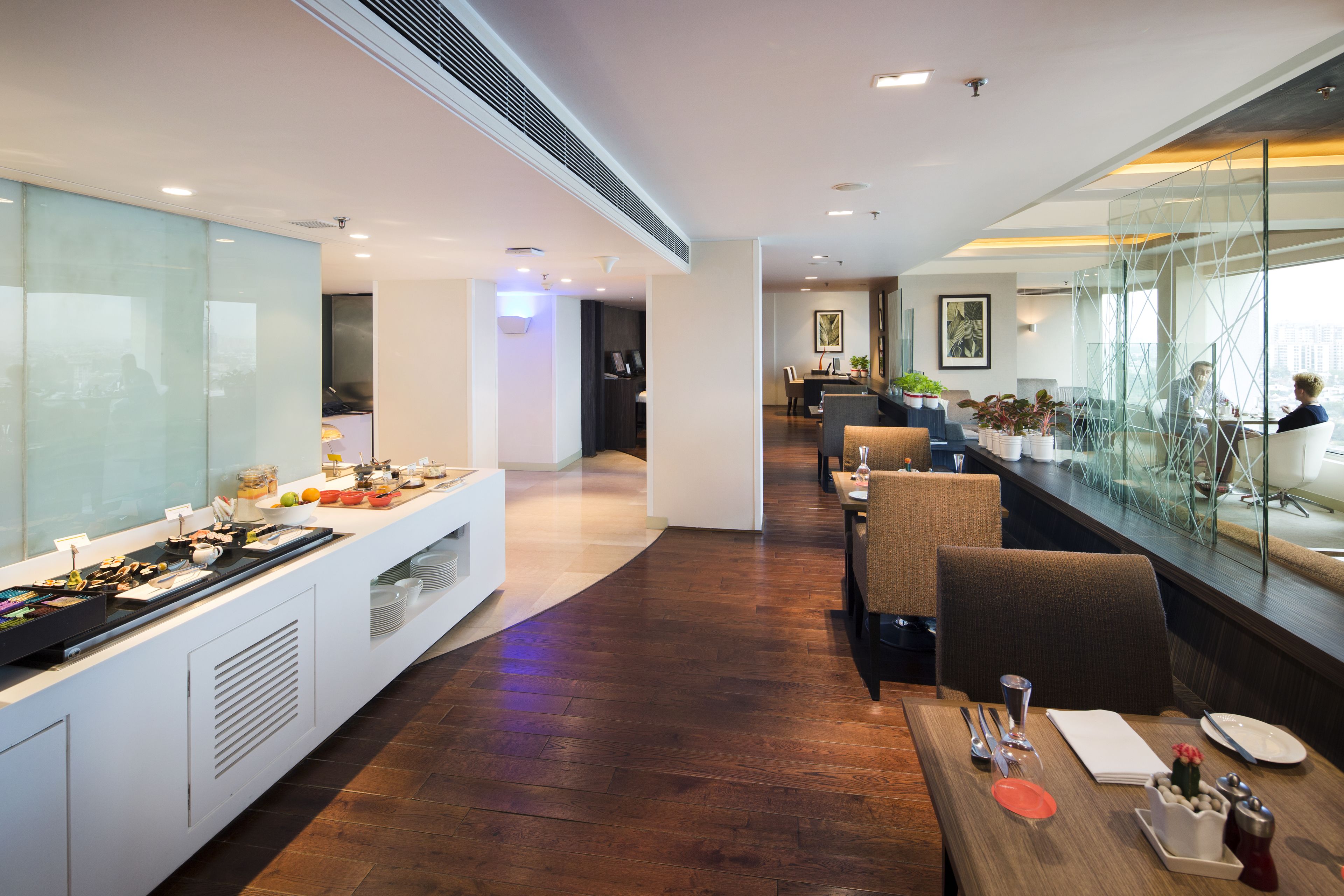 A modern hotel dining area with a buffet, seating arrangements, and a view through a glass partition.