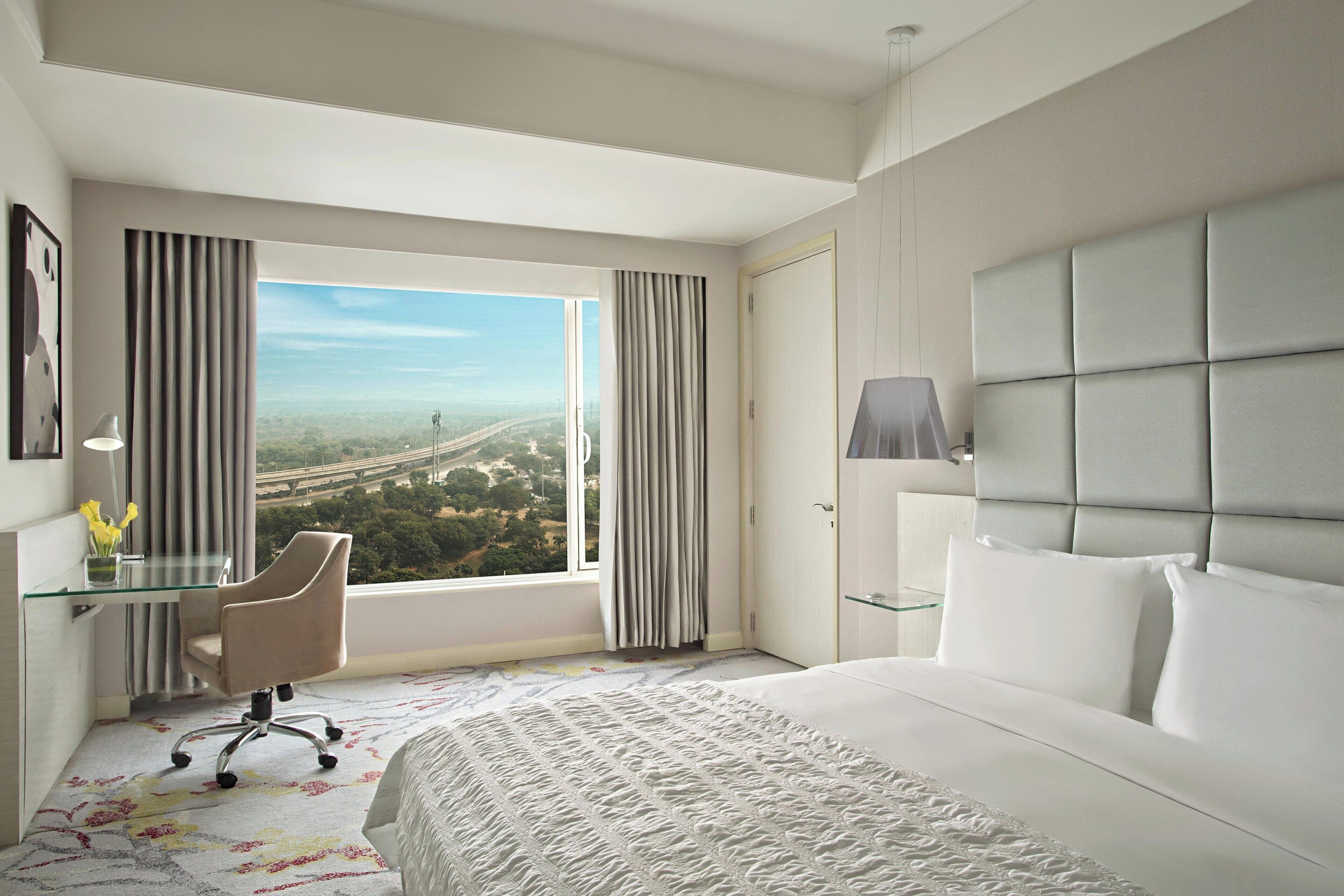 A hotel room with a large bed, a desk with a chair, a view of a cityscape, and a window with curtains.