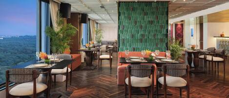 A spacious dining area with a high ceiling, large windows offering a view of the city, and a green textured wall.