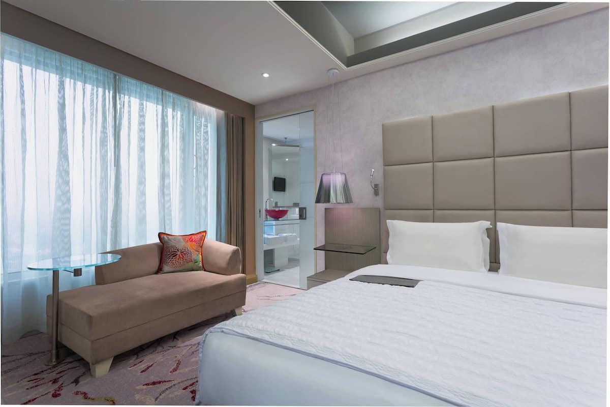 Moderni hotellihuone, jossa on suuri sänky, sohva, lasipöytä ja suuret ikkunat, joista avautuu näkymä kaupunkiin.