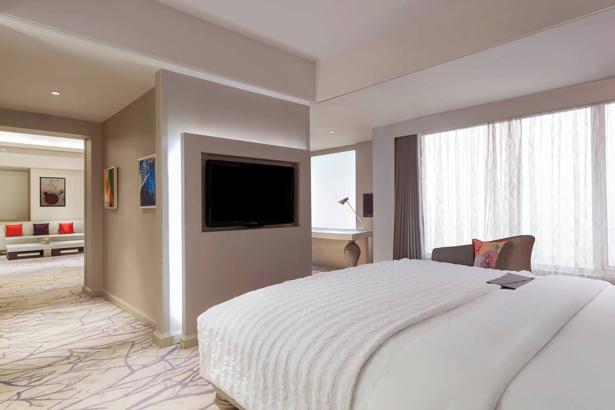 Hotellihuone, jossa on suuri sänky, seinälle asennettu taulutelevisio, työpöytä tuolilla sekä suuri ikkuna, jossa on läpikuultavat verhot.