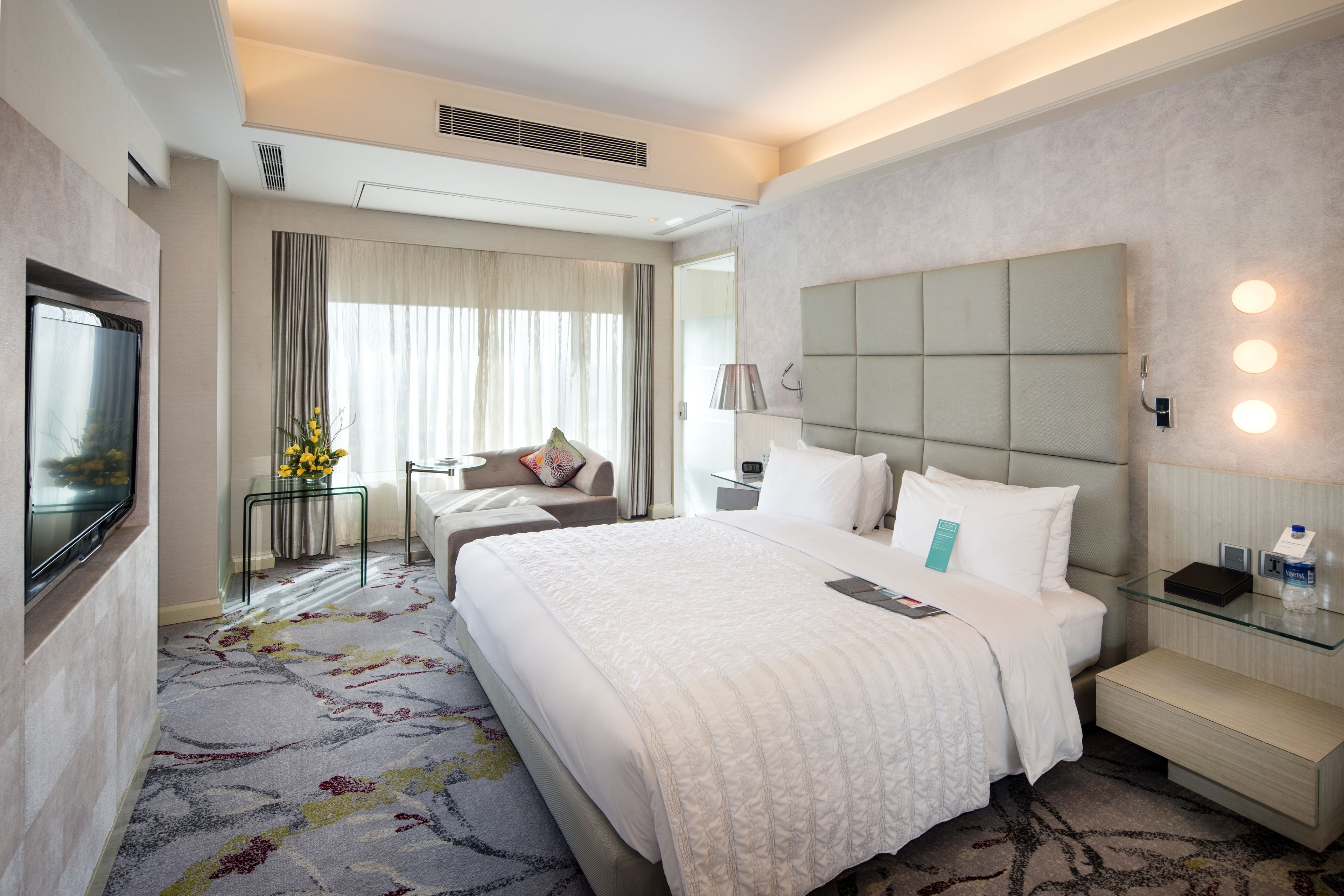 A hotel room with a large bed, a TV, a bedside table, a glass coffee table, and a window with curtains.