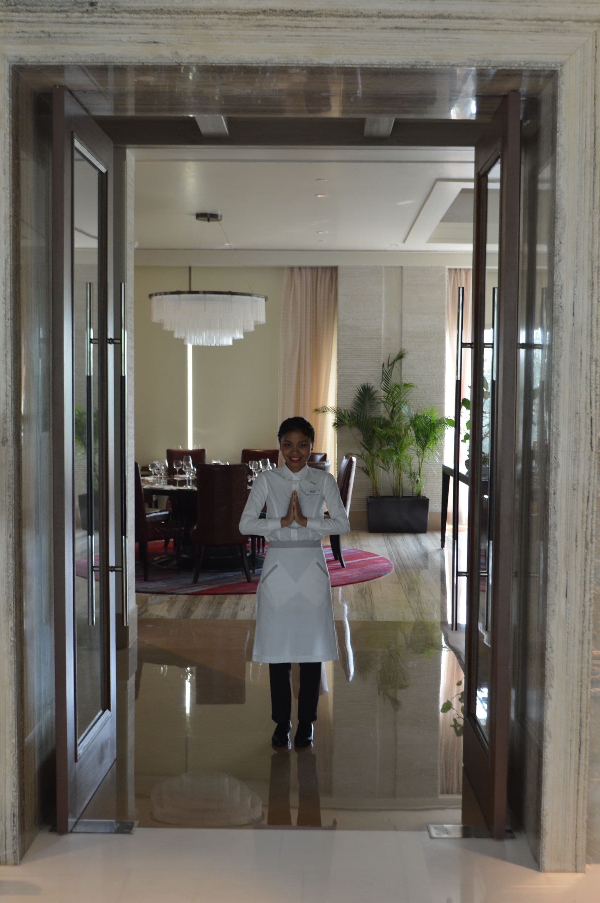 A person standing in a doorway with a polished floor, a dining area with tables and chairs in the background, and a potted plant.