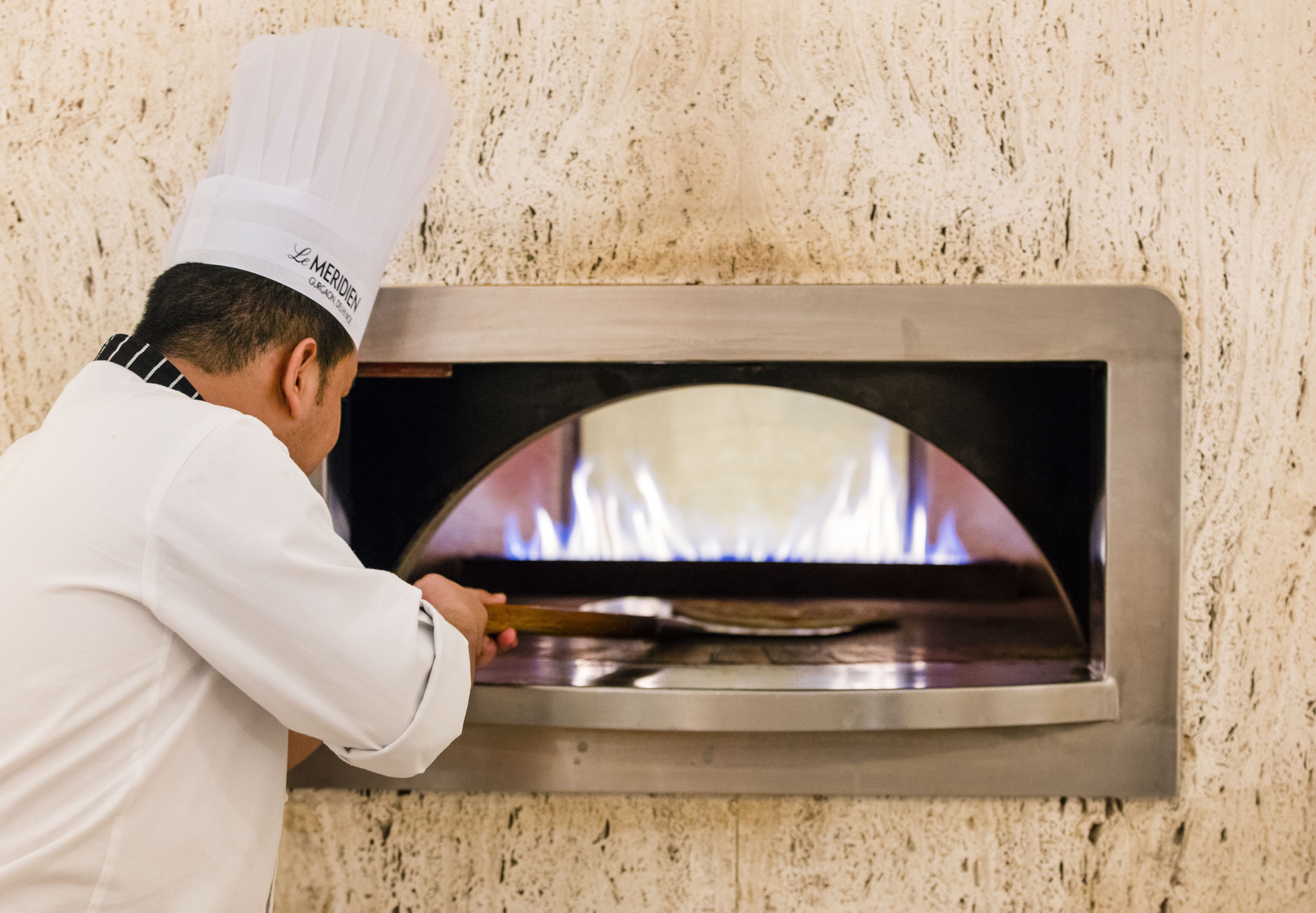 A chef in a white uniform and chef's hat operating a brick oven pizza oven.