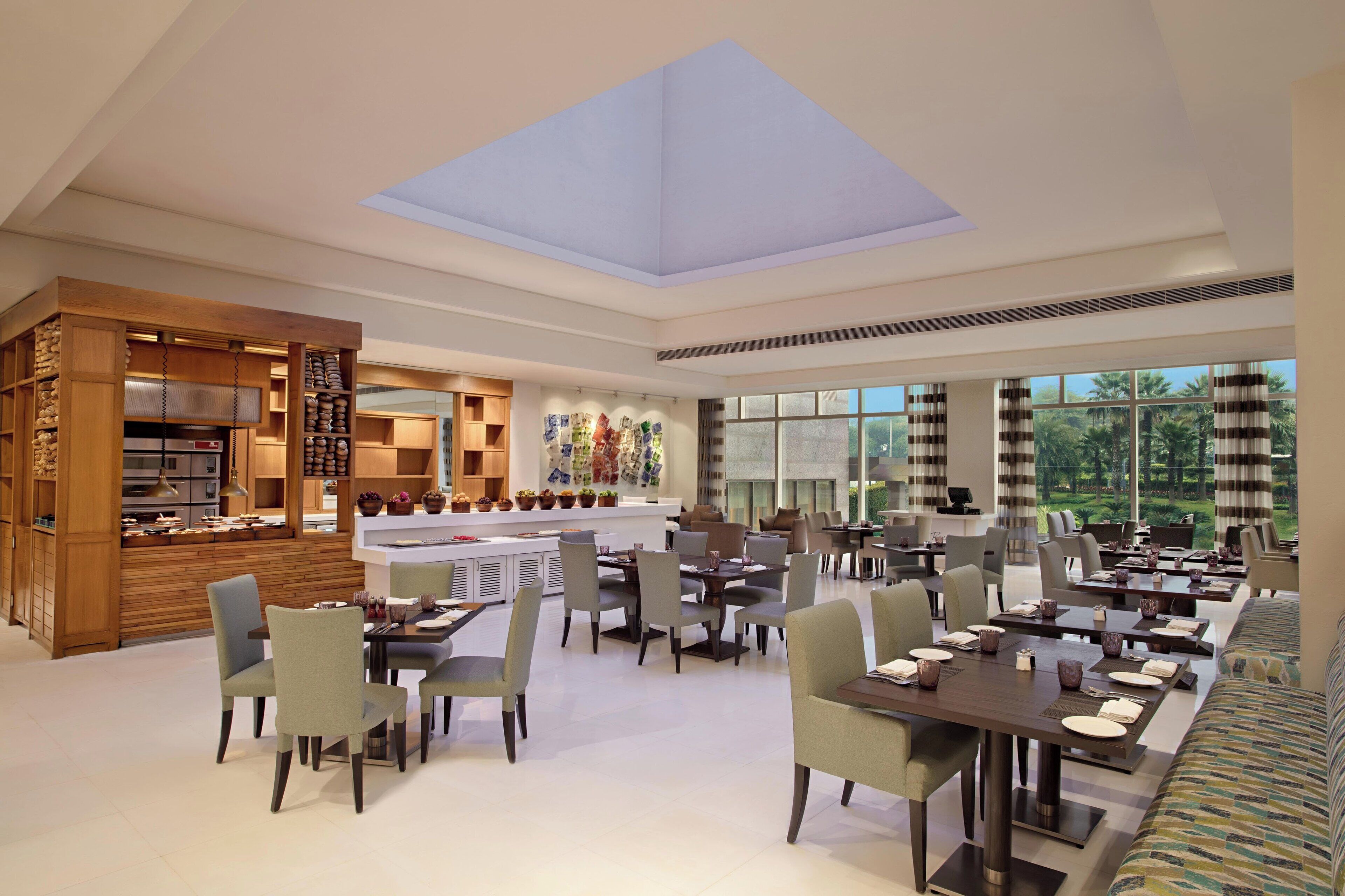 A spacious dining area with multiple tables and chairs, a kitchen area with wooden cabinets and appliances, and large windows offering a view of greenery outside.