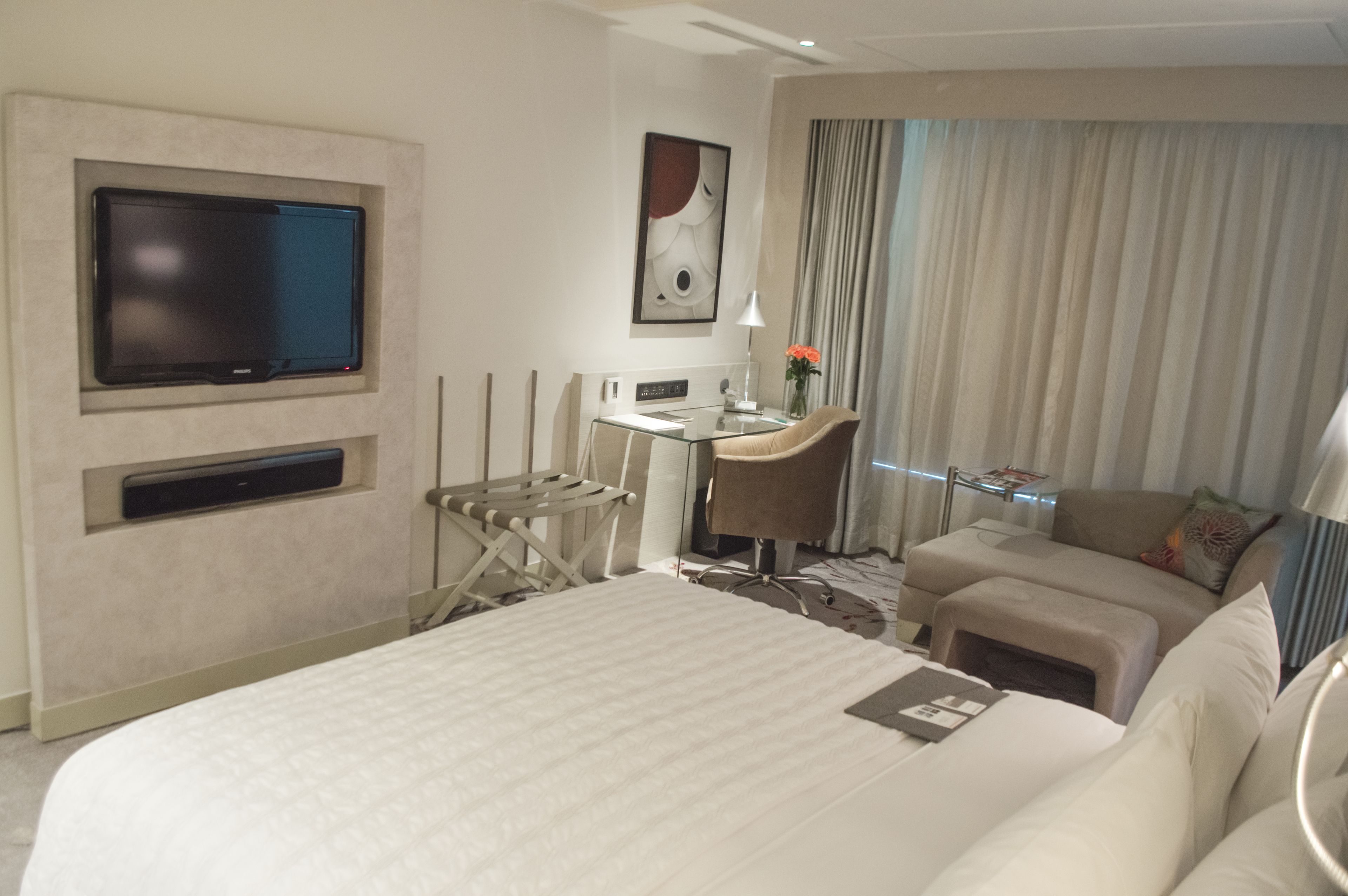 Habitación de hotel con una cama grande, un escritorio con silla, un televisor montado en la pared y una ventana con cortinas.