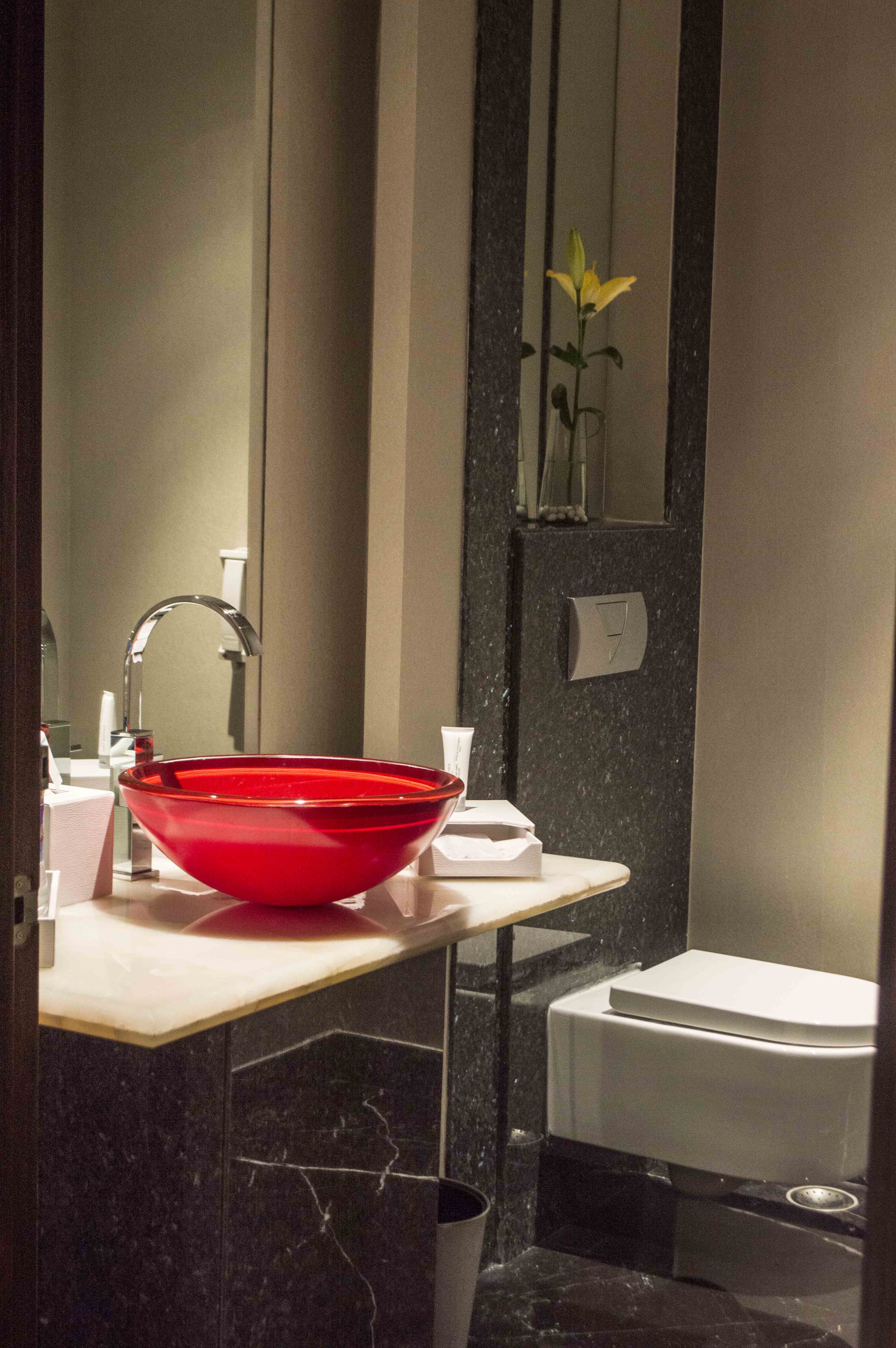 Ein modernes Badezimmer mit einem roten Waschbecken aus Glas, einer weißen Toilette und einem Spiegel, der eine gelbe Blume reflektiert.