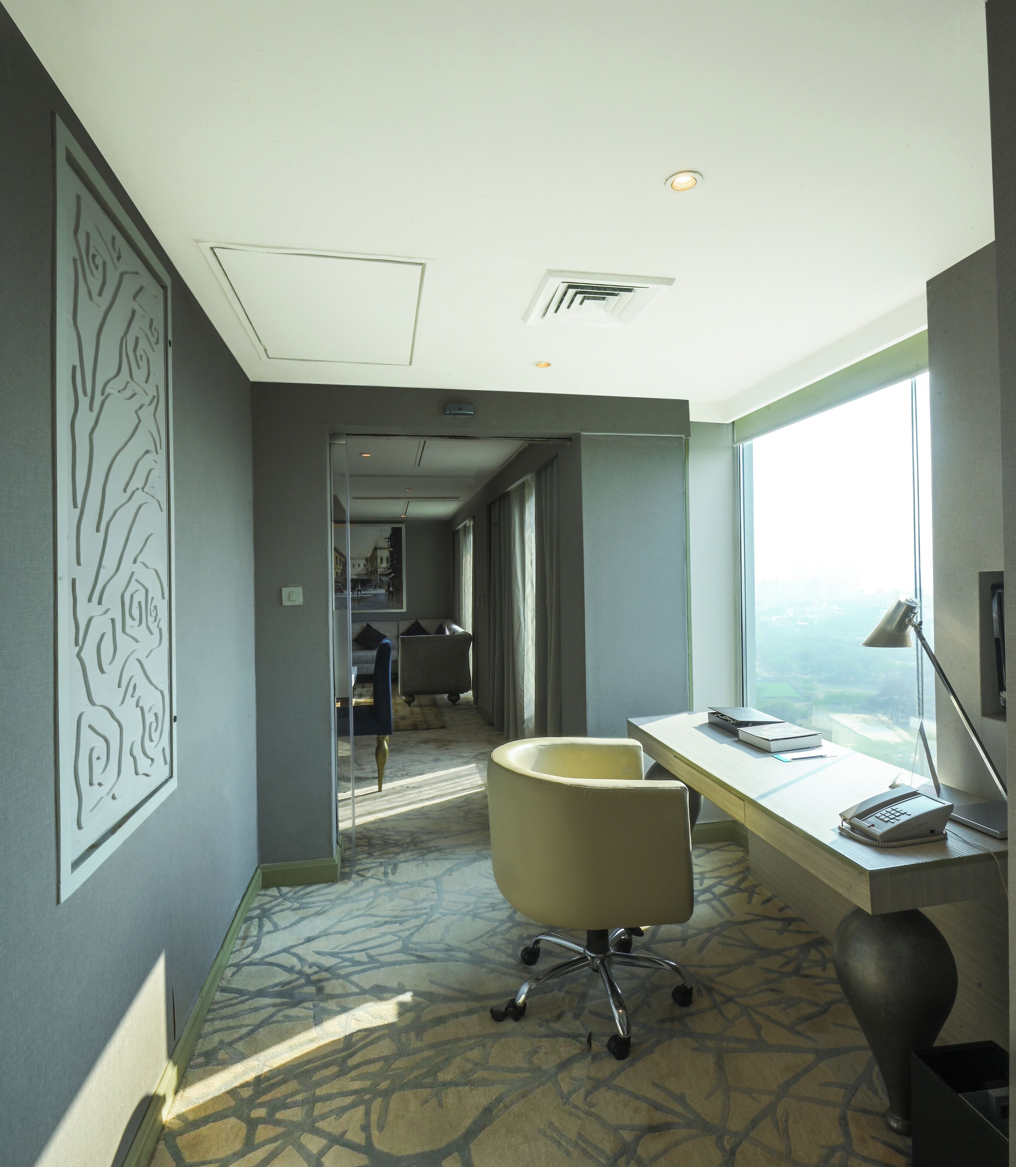 A hotel room with a large window, a desk with a lamp, a chair, and a bathroom visible in the background.