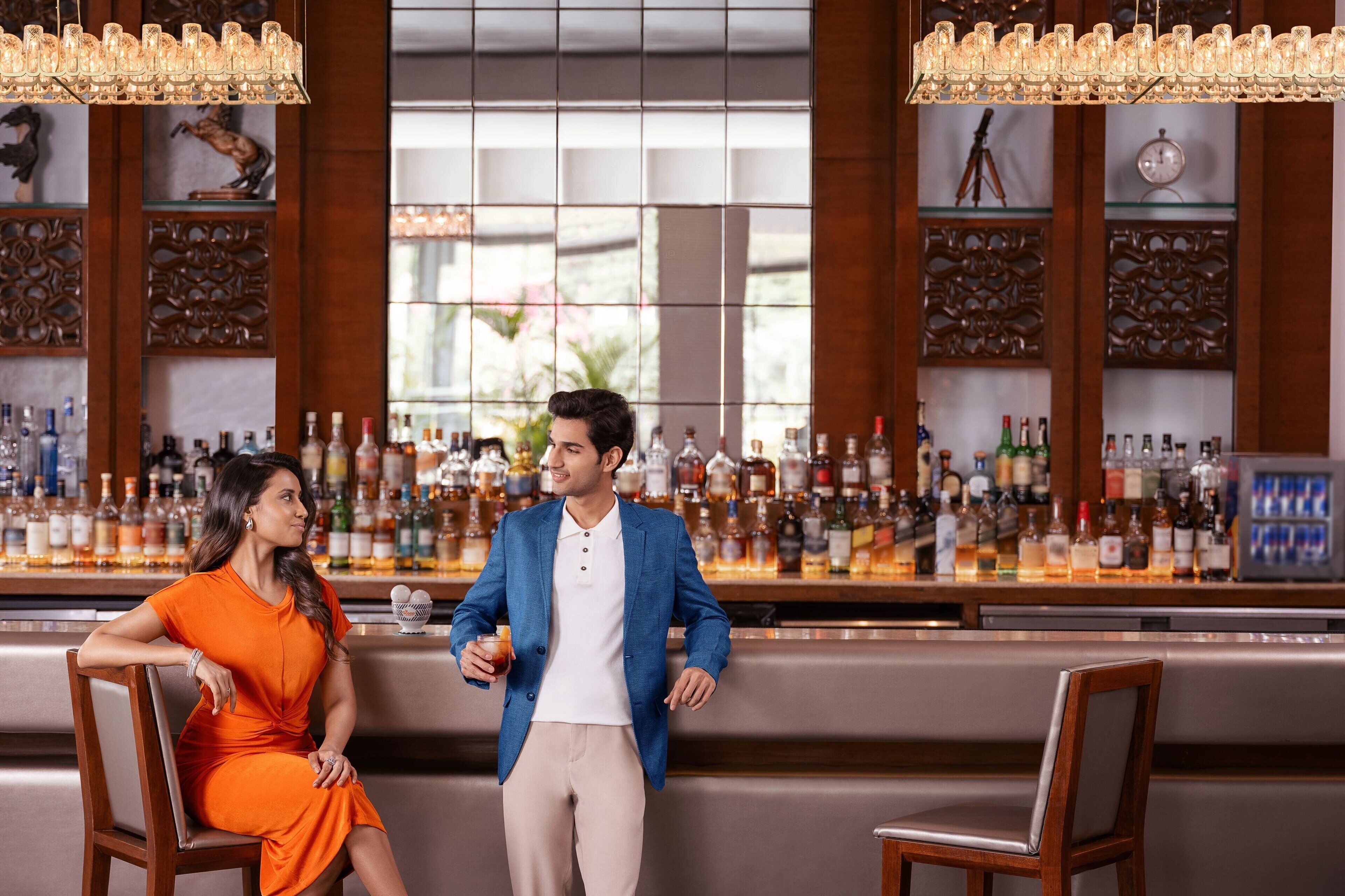 A bar area with a bartender and a woman seated. The bar has a variety of bottles and a large window in the background.