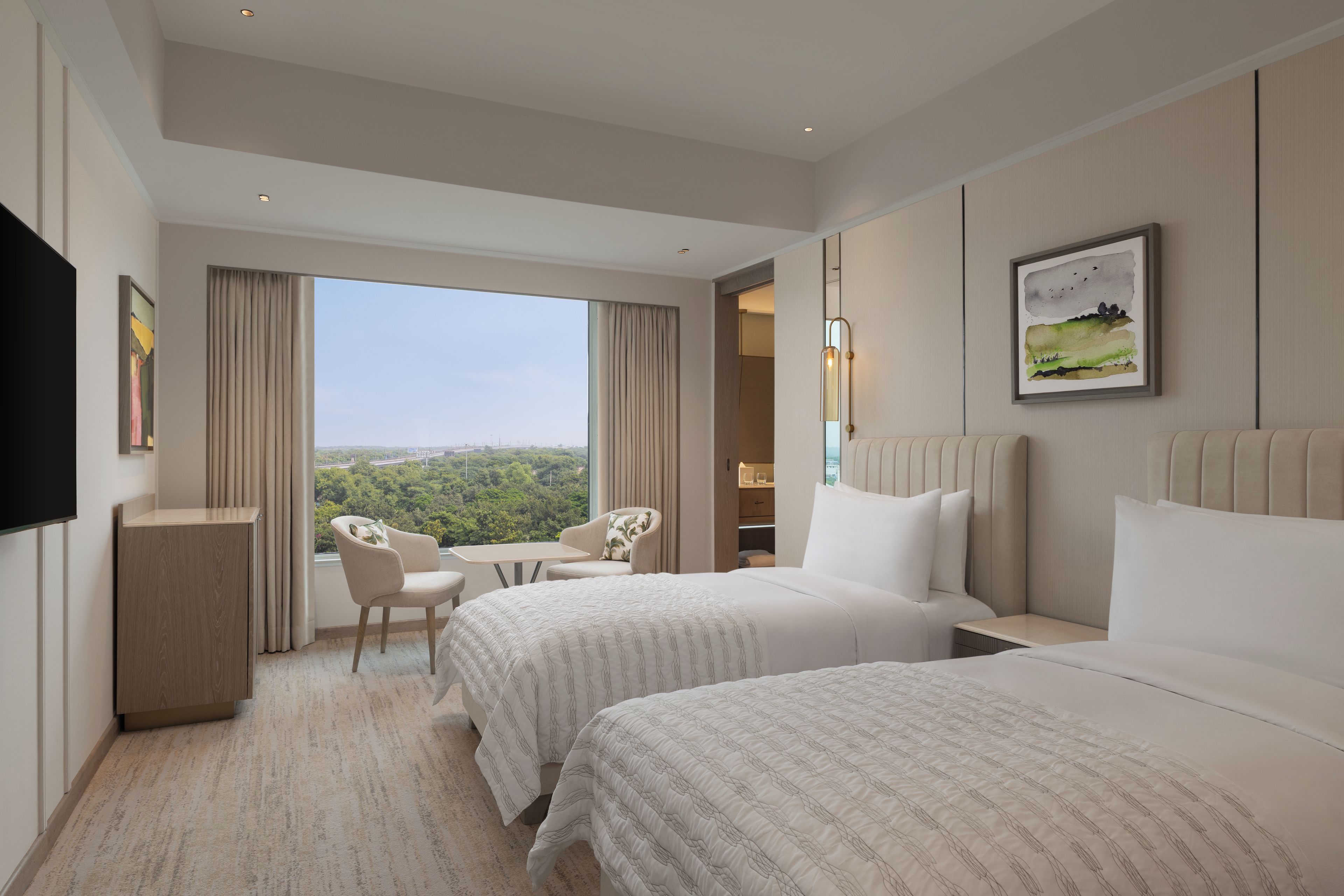 A hotel room with two beds, a desk, a chair, and a large window offering a city view.