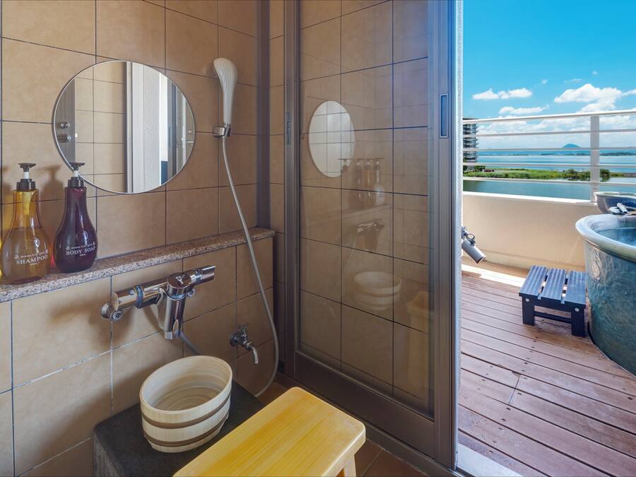 A bathroom with a shower, a sink, and a mirror. There are bottles of shampoo and body soap on the counter. The bathroom has a view of the ocean and a balcony.