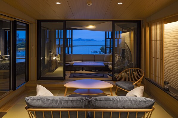 A modern interior with a glass door leading to a balcony overlooking a body of water and mountains. Inside, there is a round table with two chairs and a sofa with cushions. - Biwako Ryokusuitei (Otsu)