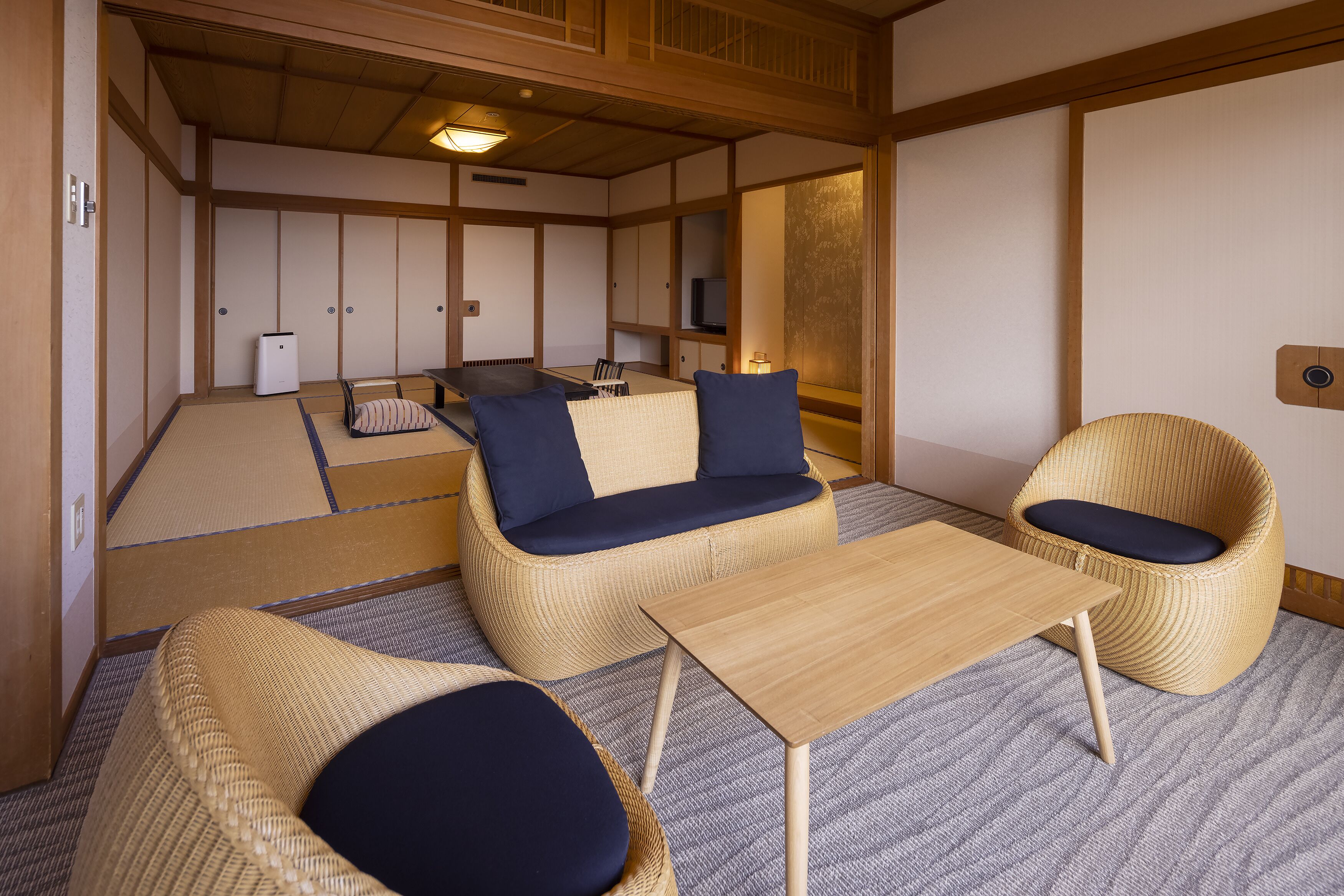 Una habitación japonesa tradicional con suelo de tatami, una mesa baja, dos sillas tejidas con cojines y un sofá con cojines.