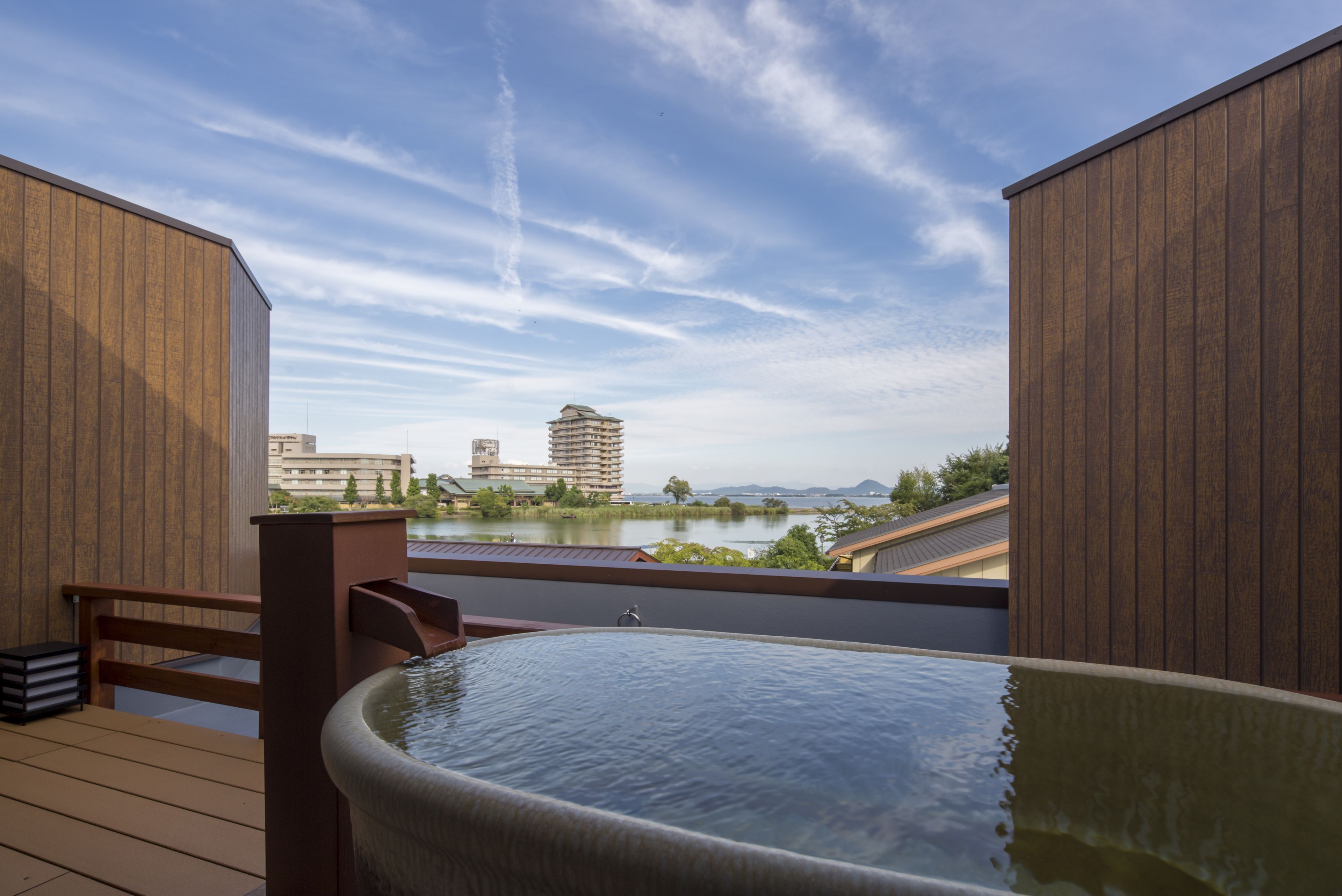 Un jacuzzi en una terraza de madera con vista a un lago y edificios al fondo.