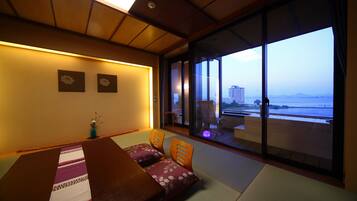 A room with tatami mats, a low table, and cushions. It has a sliding glass door leading to a balcony with a view of the sea and buildings.