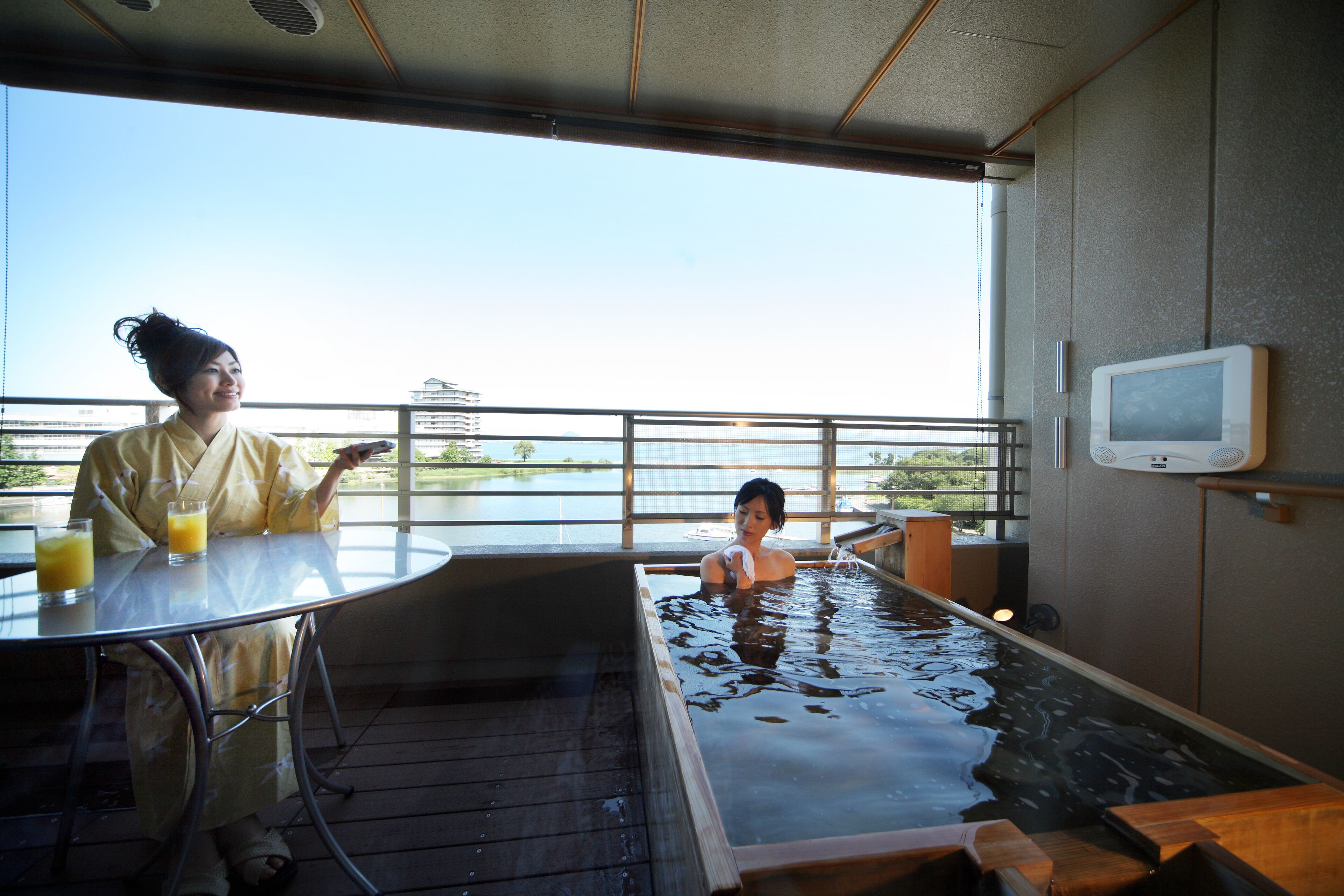 A room with a glass table, two glasses of orange juice, a wooden bathtub, and a television on the wall.