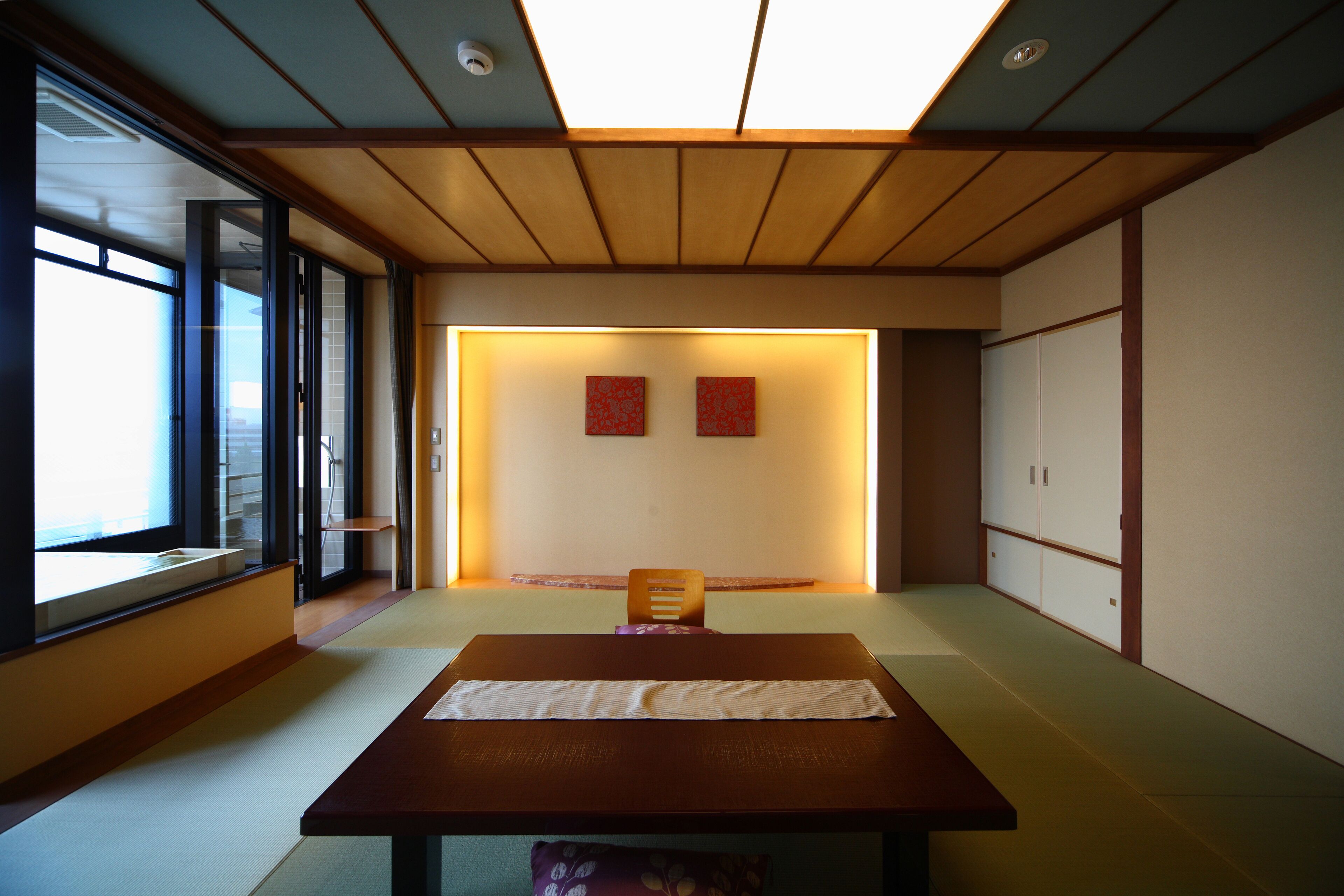 A traditional Japanese room with tatami flooring, a low wooden table, sliding doors, and a view through a large window.