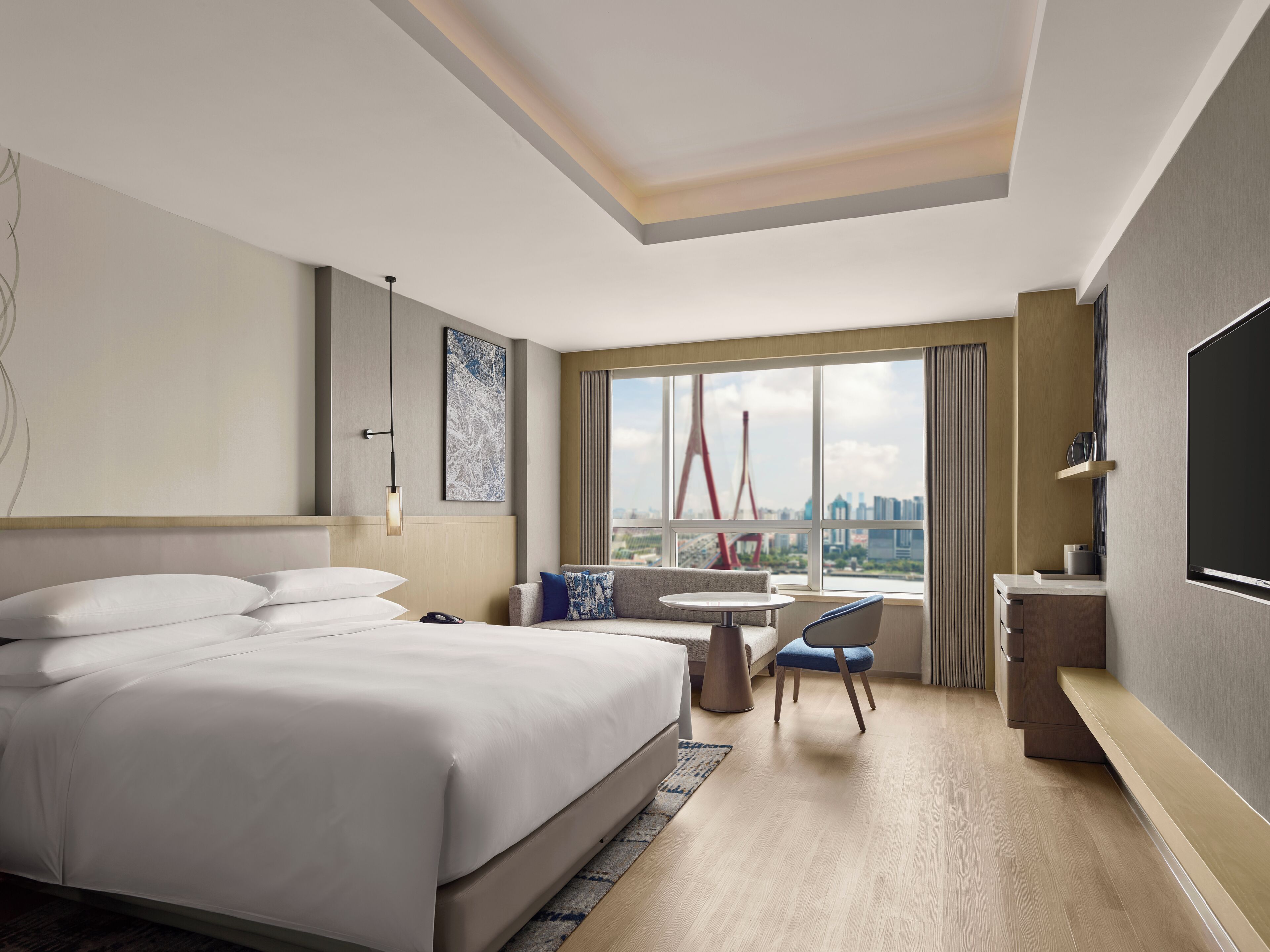 A hotel room with a bed, a desk, a chair, a TV, and a large window with a city view.