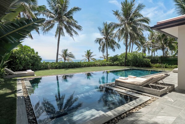 A swimming pool with lounge chairs, surrounded by palm trees and a view of the ocean.