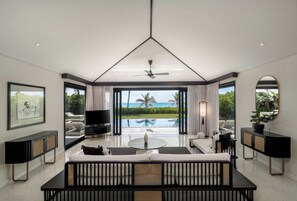 A modern living room with a large TV, a sofa, a coffee table, and a view of a pool and palm trees. - TIA Wellness Resort - Spa Inclusive (Da Nang)