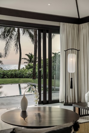 A modern room with a large window, a round table, and a view of a palm tree and water body. - TIA Wellness Resort - Spa Inclusive (Da Nang)