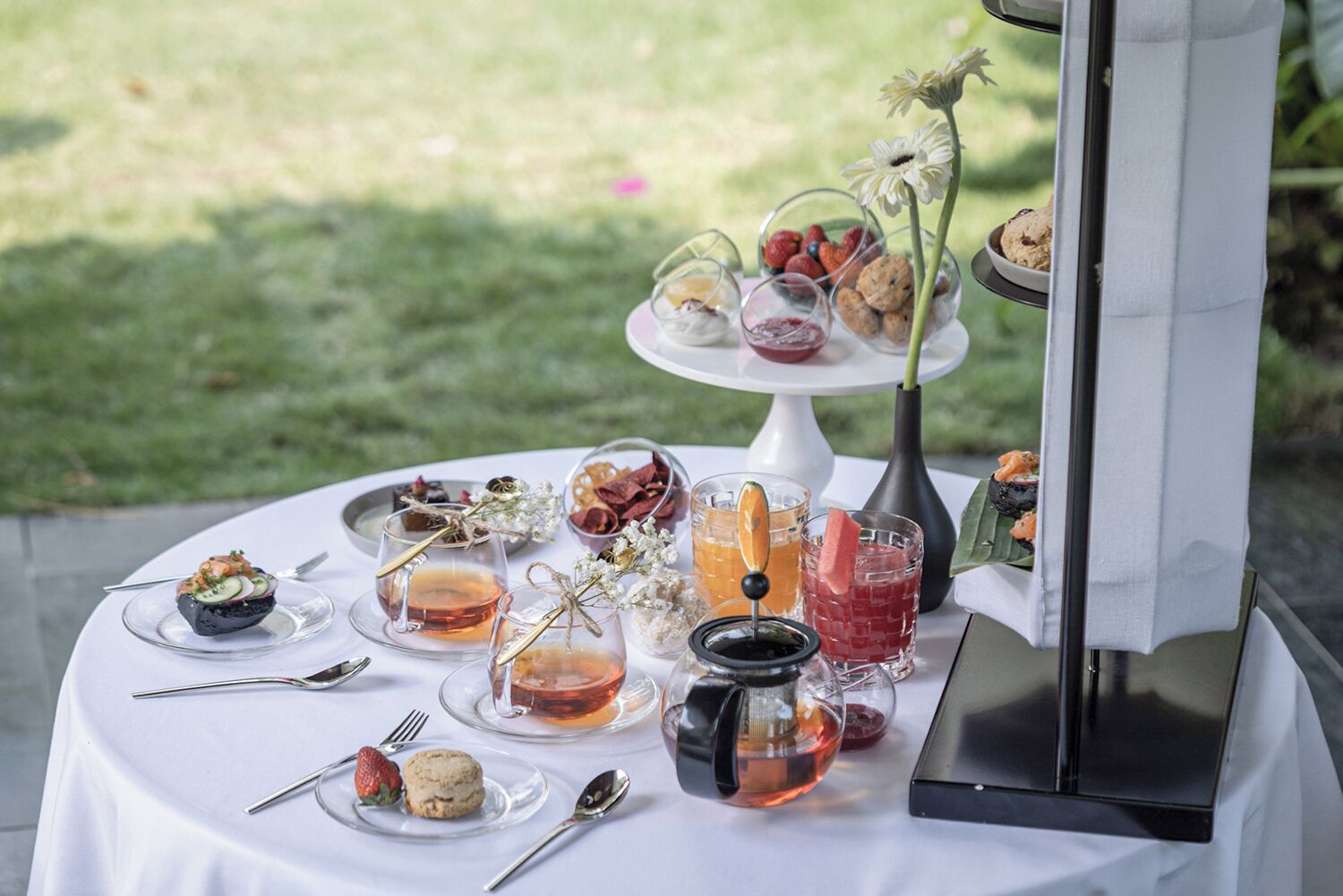 A table set outdoors with various food items, drinks, and a small decorative flower arrangement.