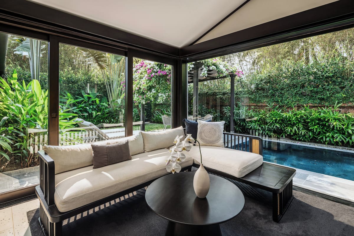 A modern outdoor seating area with a sofa and coffee table, a pool, and lush greenery visible through large windows.