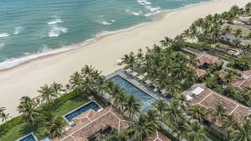 An aerial view of a resort with a swimming pool, palm trees, and a beach.