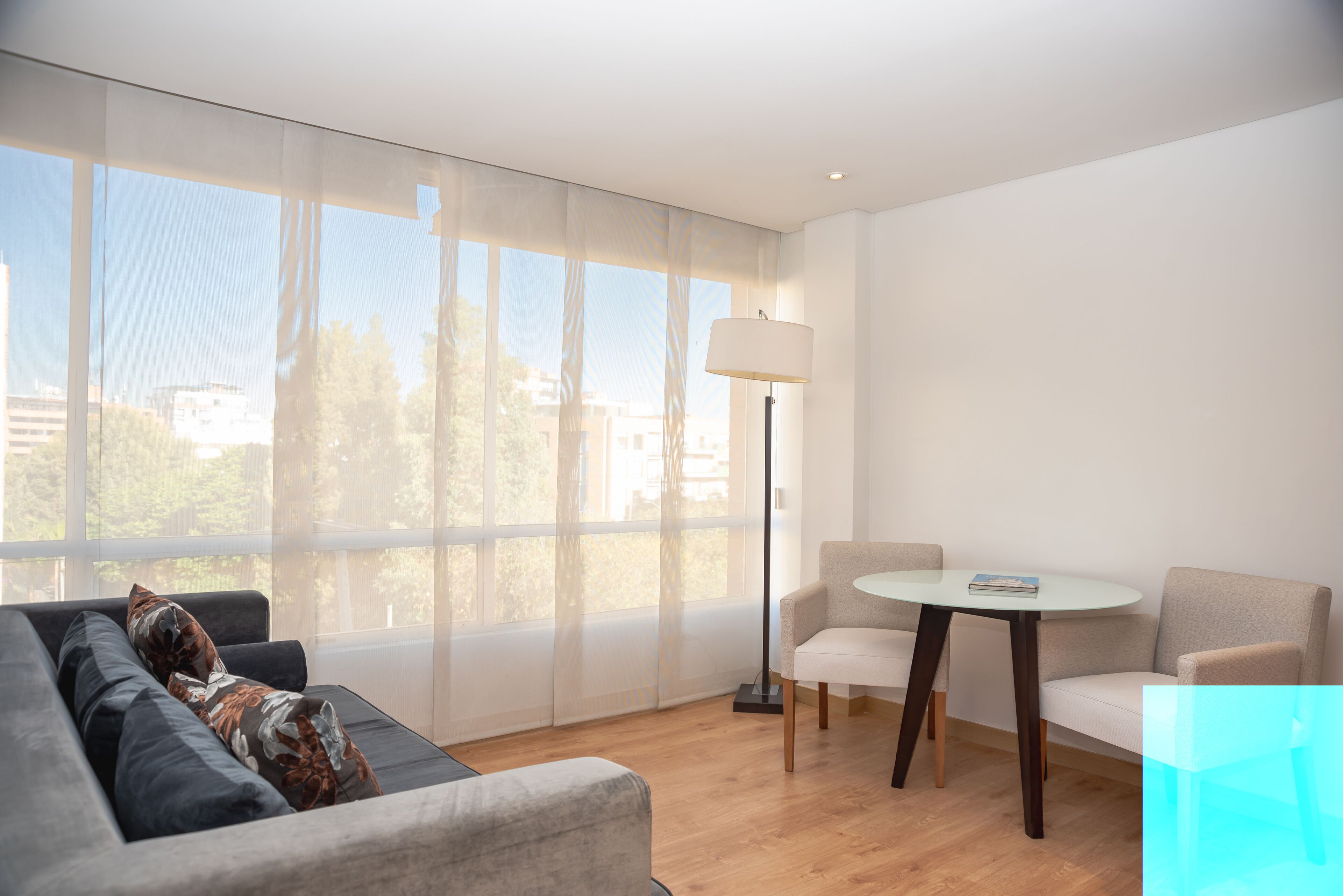 Una sala moderna con un sofá, dos sillones, una mesita y una lámpara de pie. Amplias ventanas ofrecen vistas al exterior.