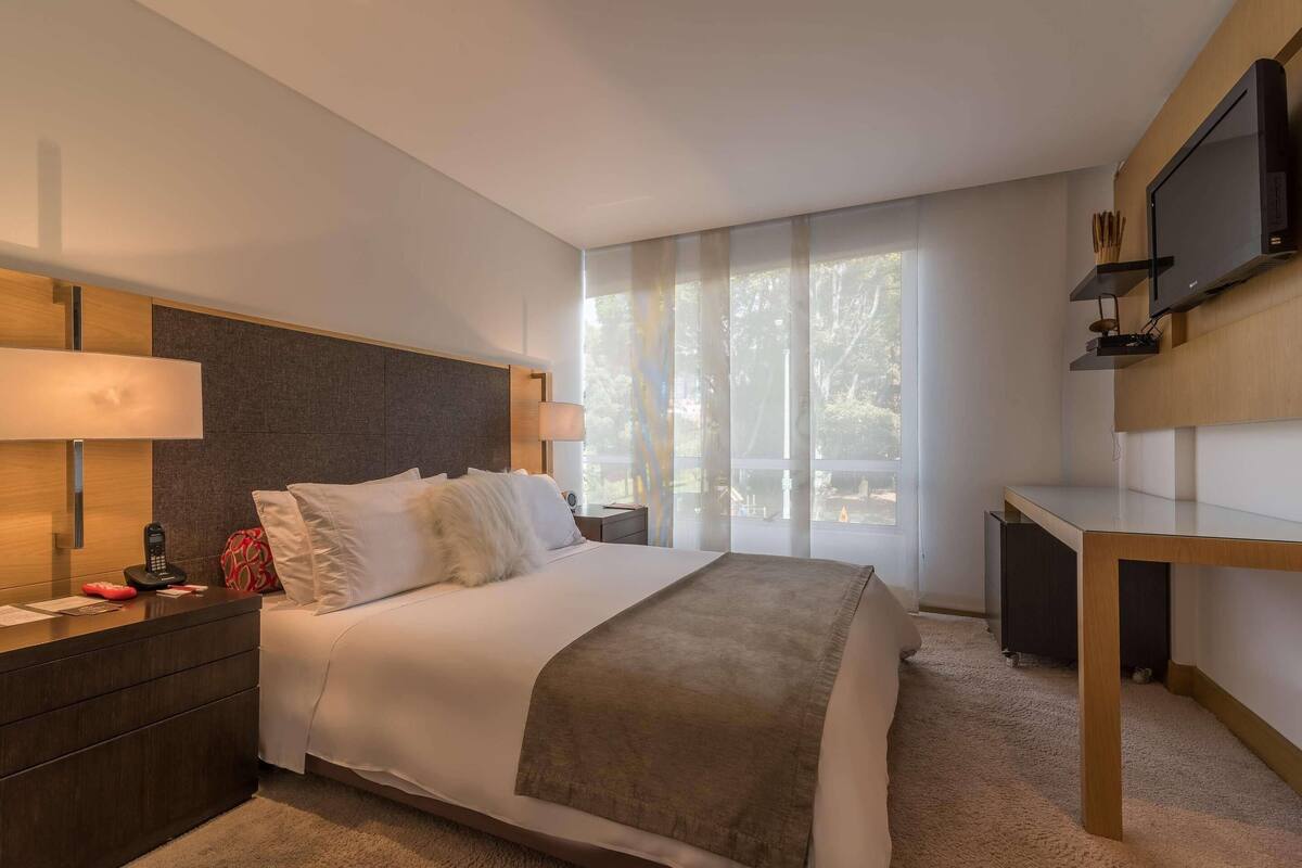 Quarto de hotel moderno com cama grande, mesas de cabeceira, escrivaninha, televisão e janelas amplas com cortinas translúcidas.