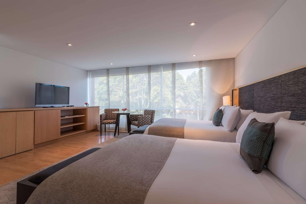 Quarto de hotel moderno com uma cama grande, uma escrivaninha com televisão, uma mesinha com cadeiras e amplas janelas com vista para árvores.