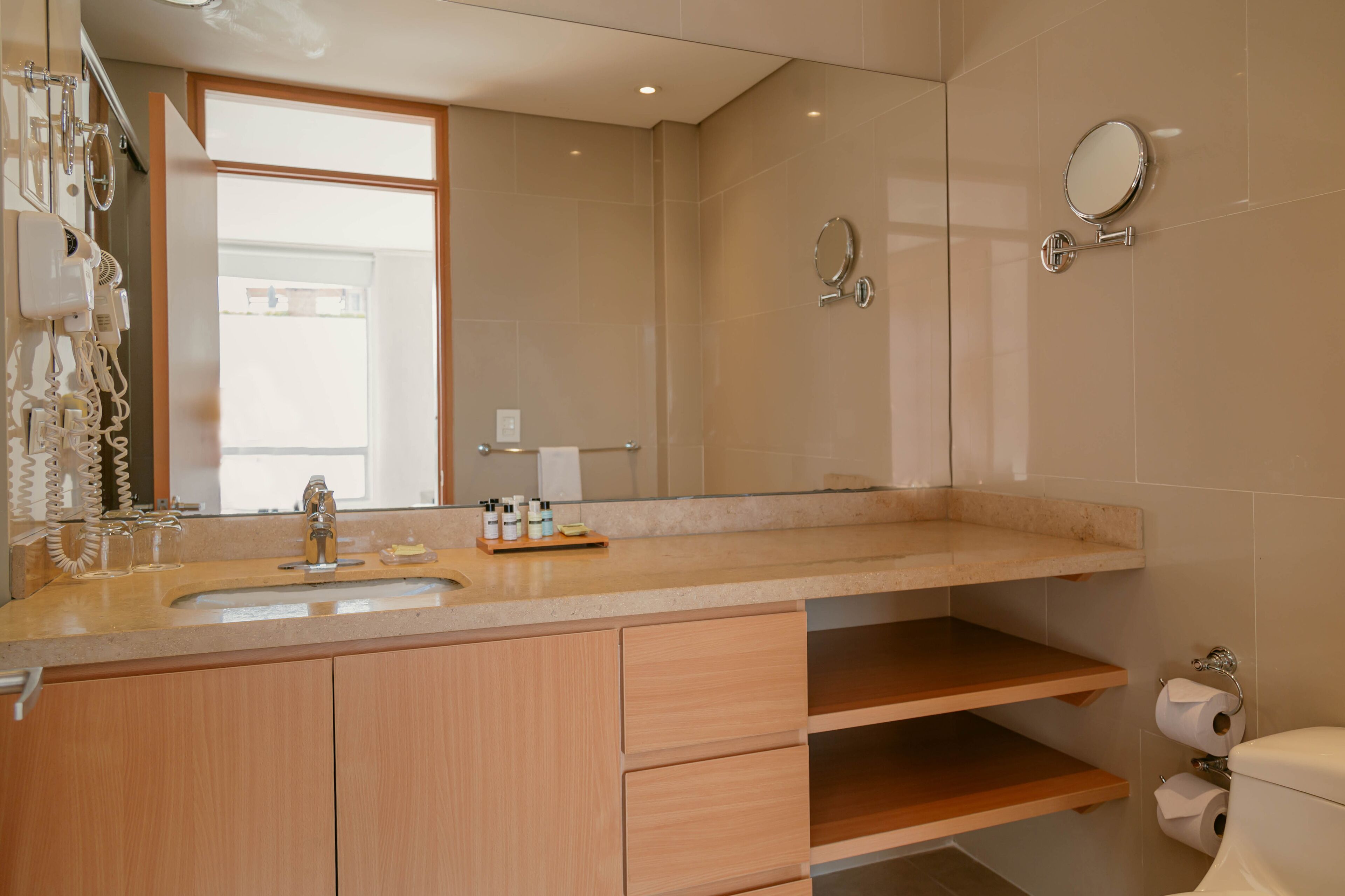 Un baño moderno con un espejo grande, un lavamanos y un inodoro. Hay armarios de madera y un estante con artículos de aseo.