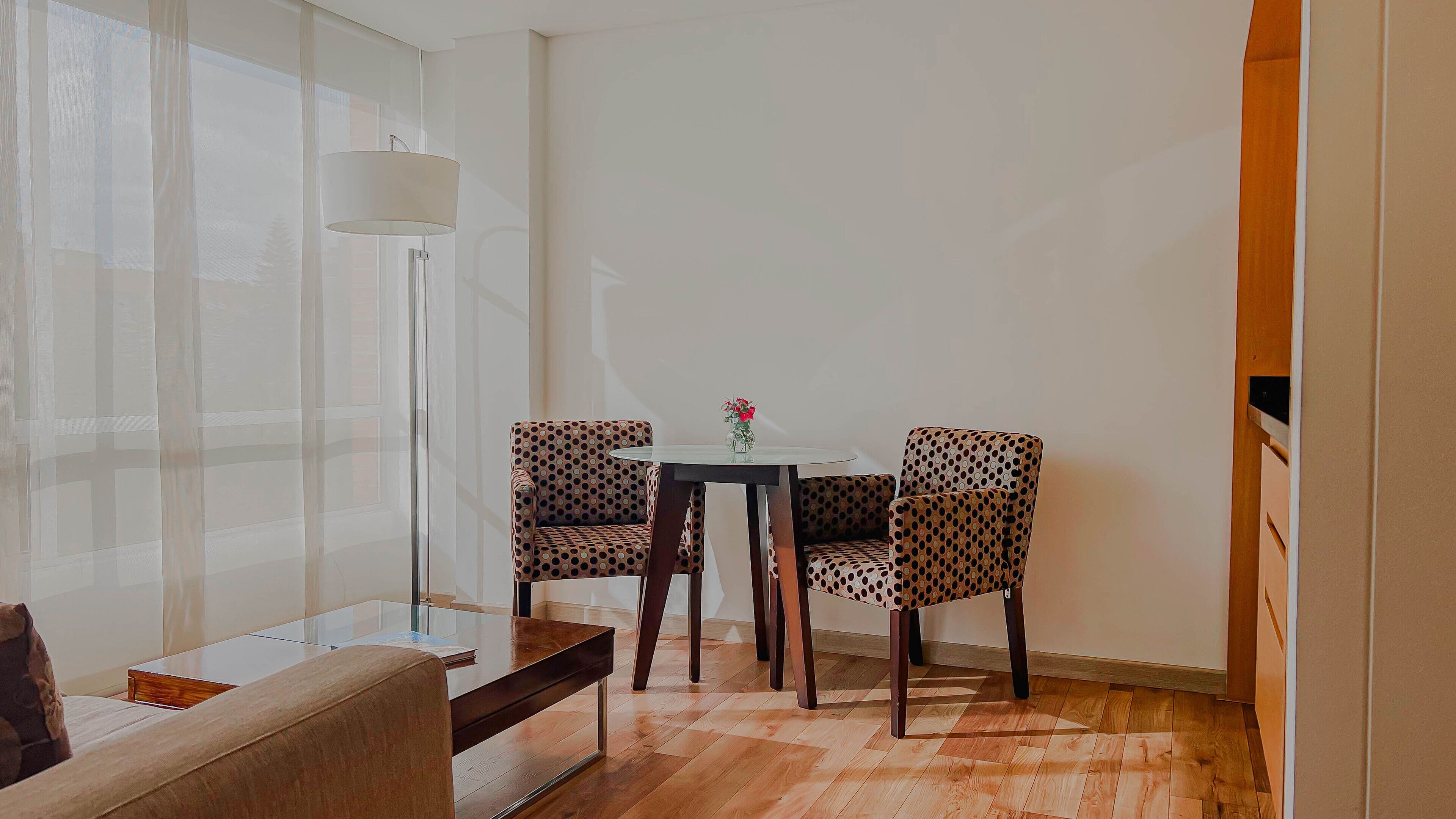A small, well-lit room with a glass table, two patterned chairs, a floor lamp, and a wooden bench.