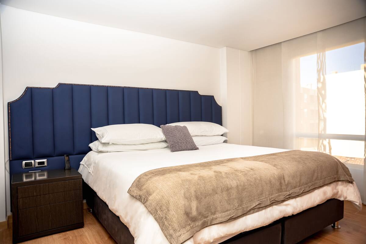 Quarto moderno com uma cama grande, cabeceira escura, mesa de cabeceira com um telefone e uma janela com cortinas.