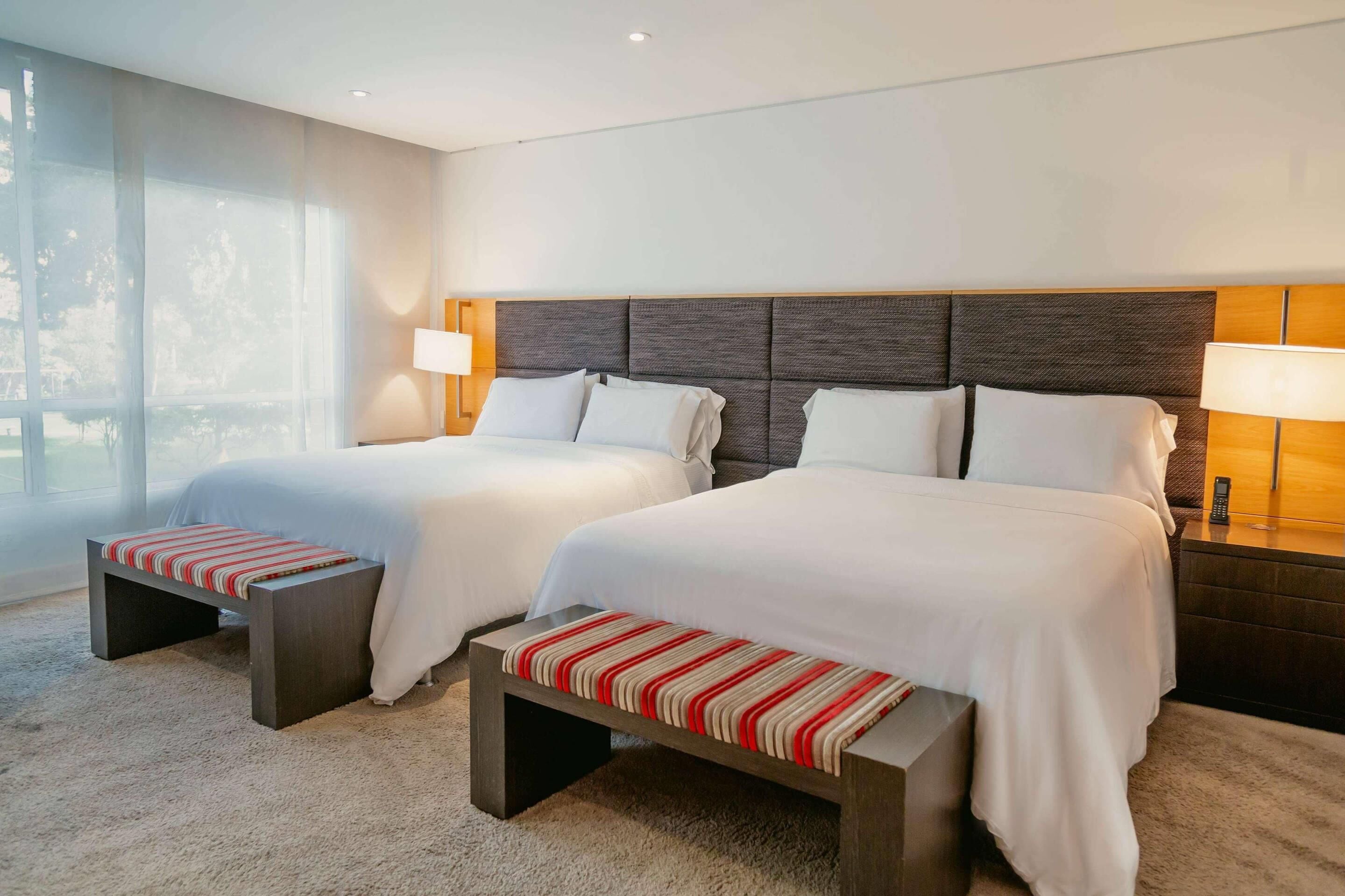Habitación de hotel con dos camas, lámparas de noche, un ventanal grande y piso alfombrado.