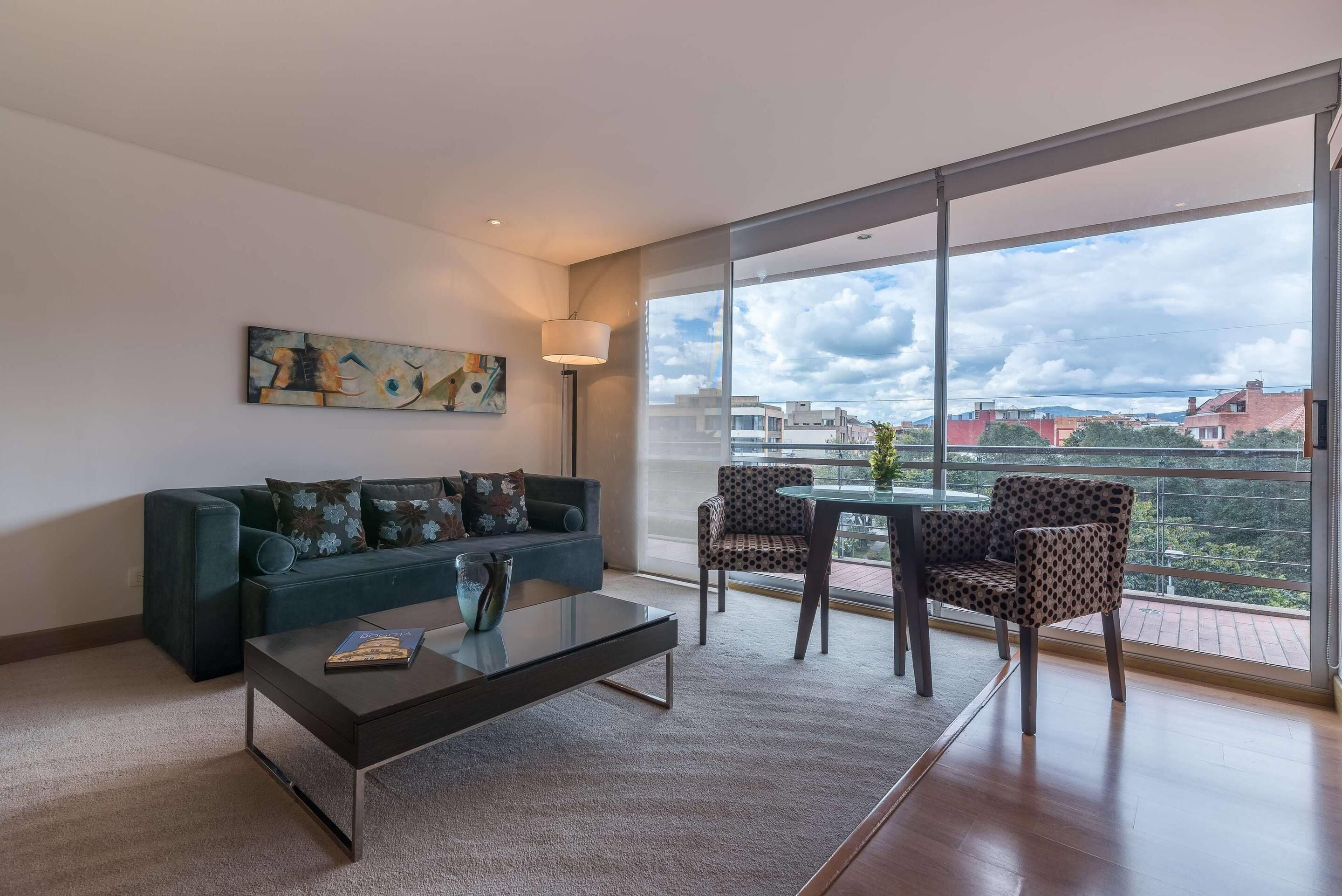 Una sala de estar moderna con sofá, mesa de centro y sillas estampadas. Un cuadro adorna la pared, y hay un ventanal con vistas a edificios y nubes.